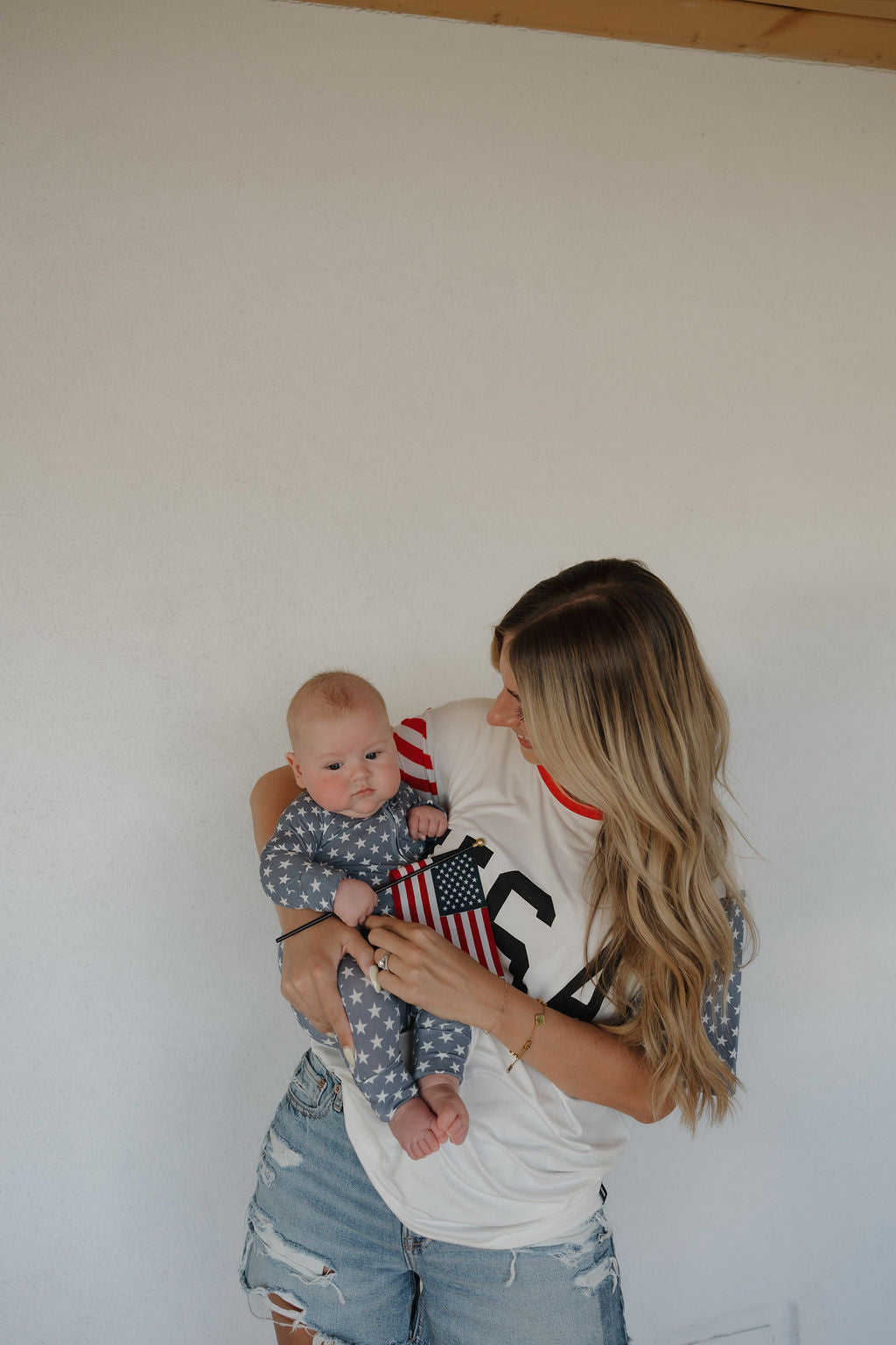 A woman wears the forever french baby Adult Bamboo T-Shirt | Stars & Stripes while holding a baby in a star-patterned, hypo-allergenic outfit. The baby waves American flags against a plain white wall.