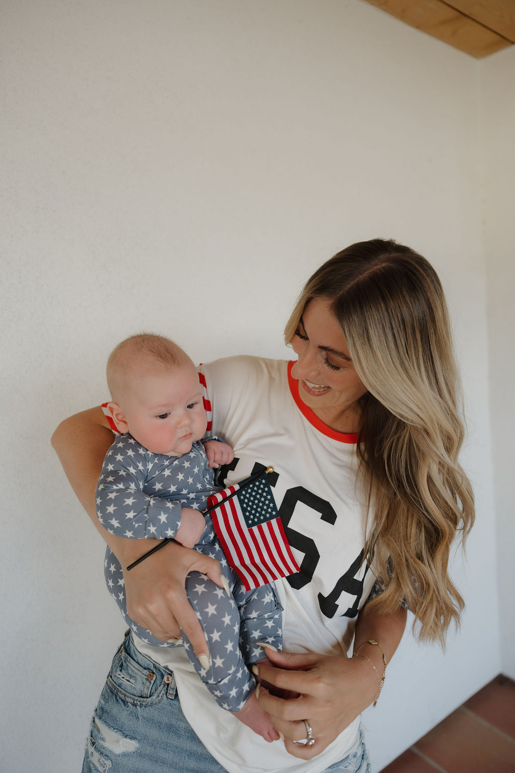 A woman wearing the forever french baby Adult Bamboo T-Shirt | Stars & Stripes smiles while holding a baby in a star-patterned, breathable outfit, as the baby waves a small American flag in a cheerful, casual setting.