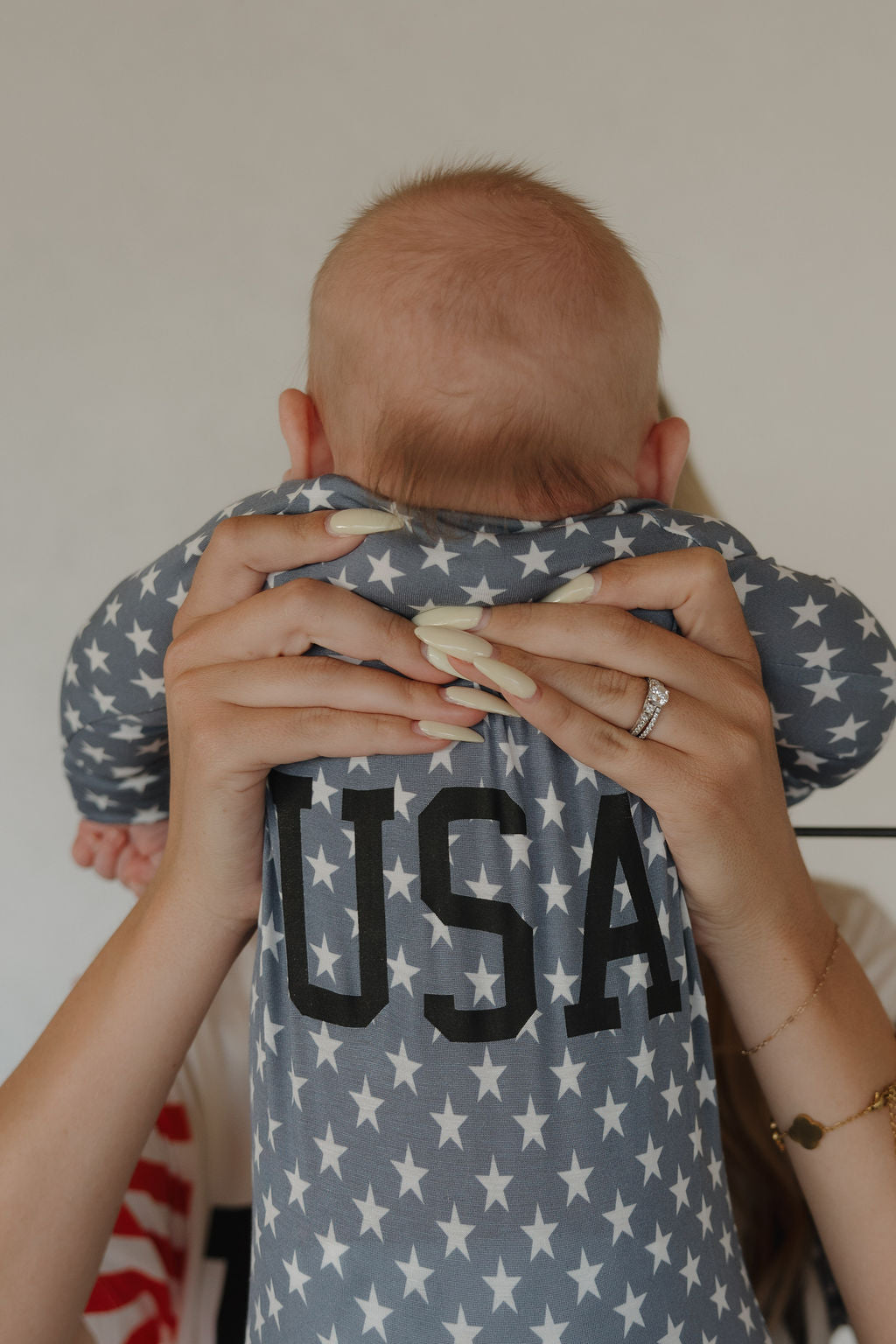 A person holds up a baby in forever french baby’s Bamboo Zip Pajamas | Star Spangled—breathable gray pajamas with white stars and "USA" on the back. The person's manicured hands and ring gently support the little one.