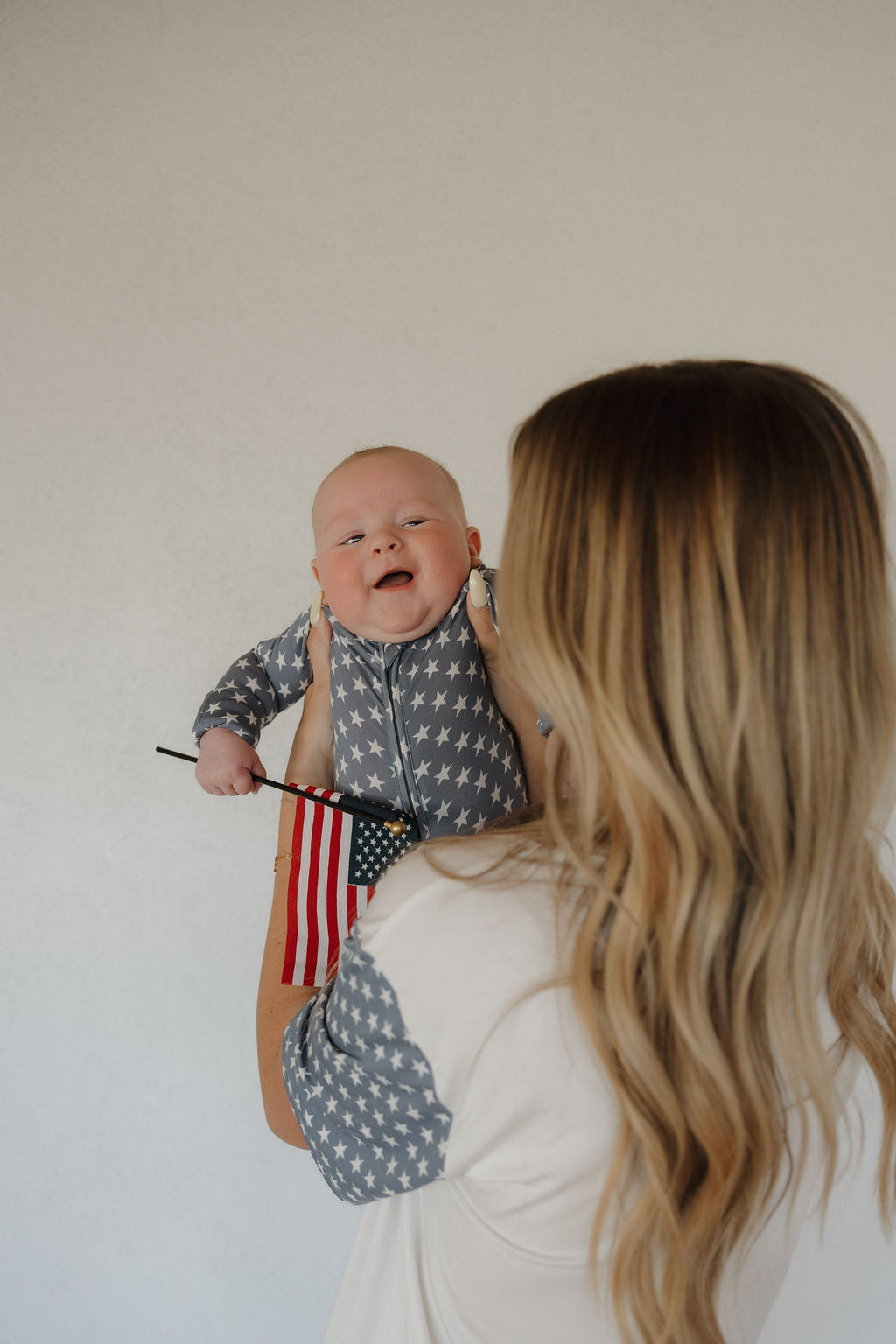 A woman with long blonde hair holds a smiling baby in forever french baby's Bamboo Zip Pajamas | Star Spangled pattern, holding a small American flag. The background is plain and light-colored.