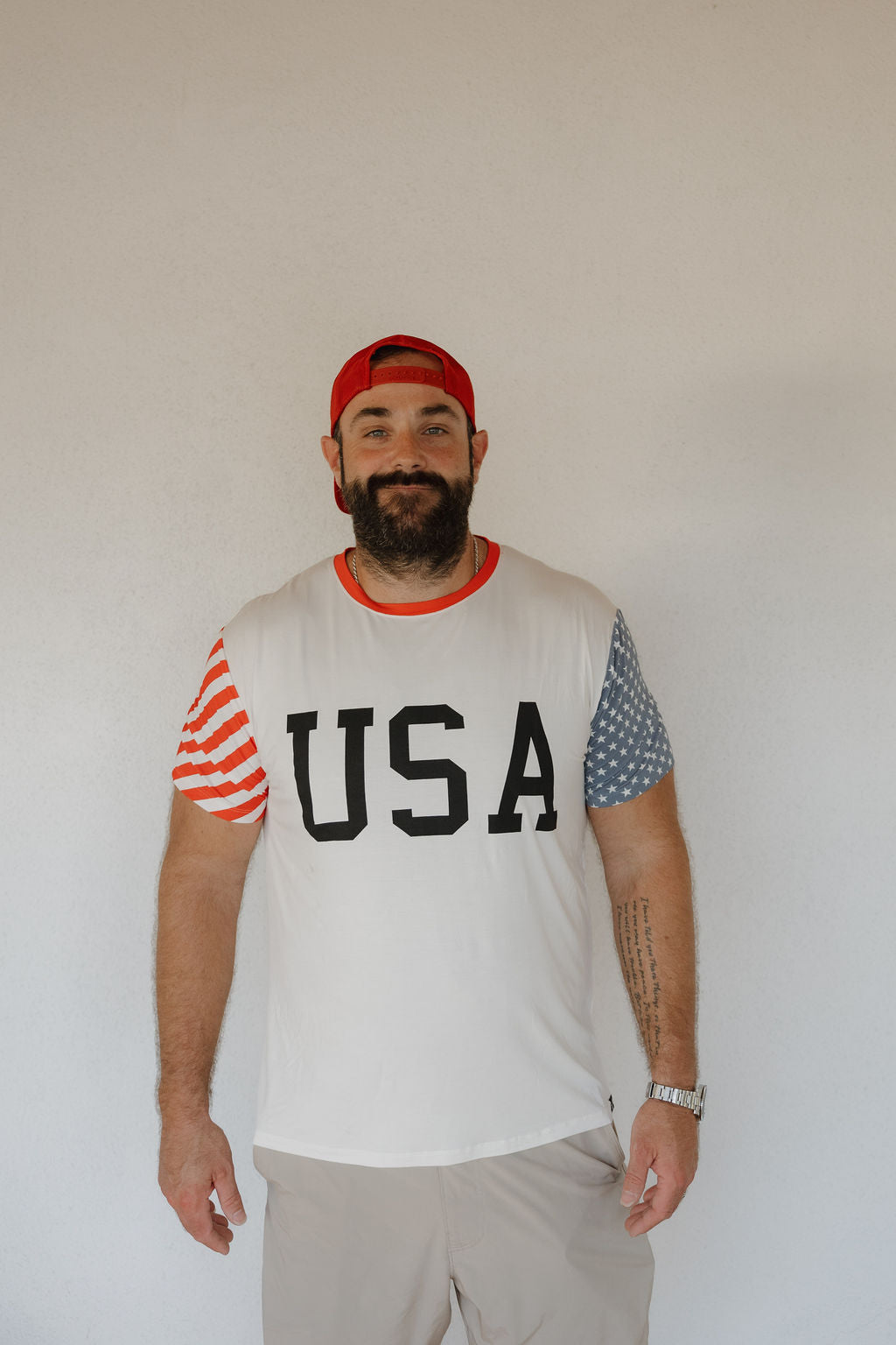 A bearded man in a red cap wears the forever french baby Adult Bamboo T-Shirt | Stars & Stripes, featuring "USA" on the front and American flag sleeves, standing against a plain light-colored wall.