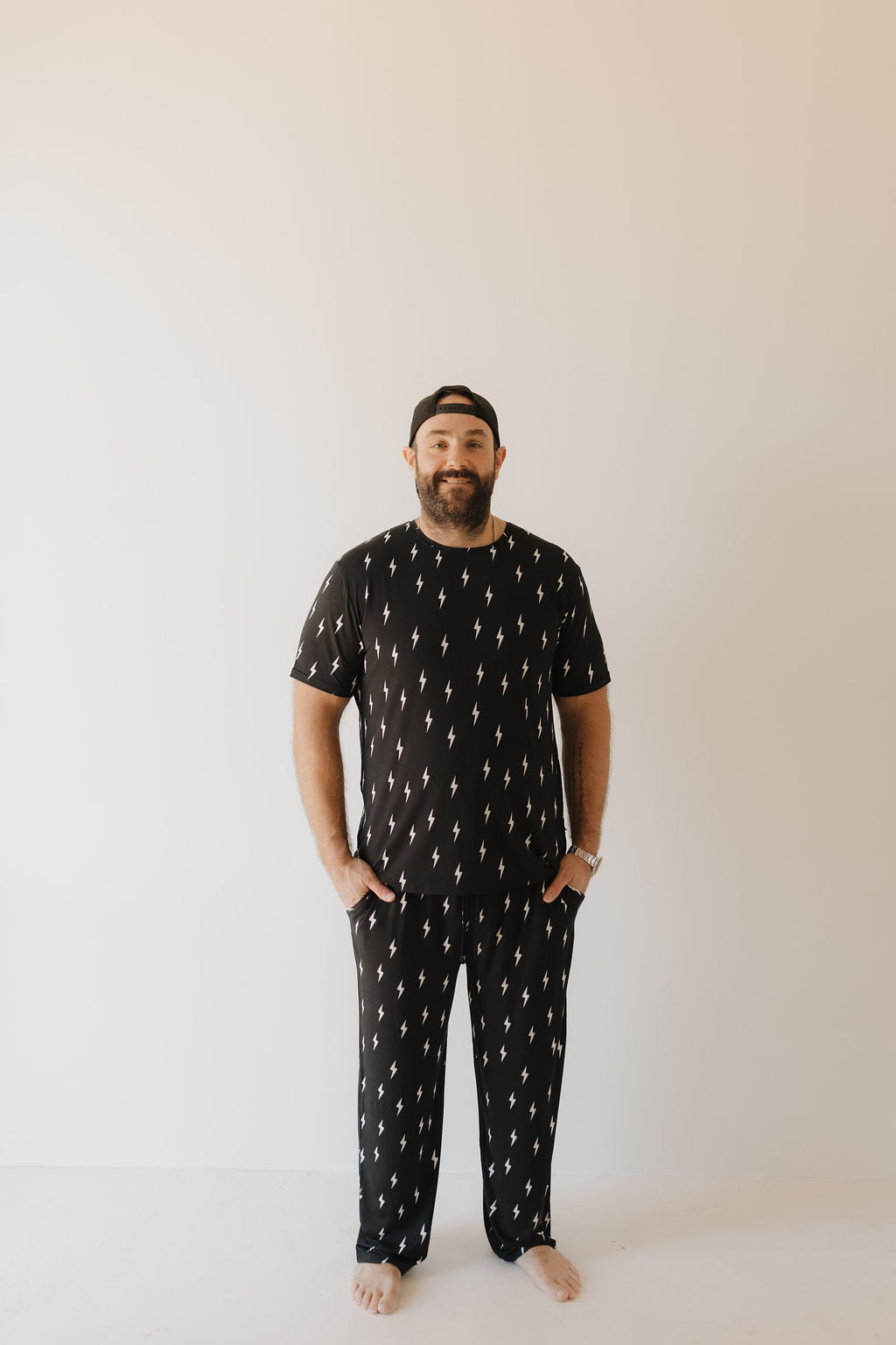 A barefoot man, with a beard and cap, stands in Men's Bamboo Short Sleeve Pajamas by Forever French Baby. Featuring midnight lightning bolt patterns on black fabric, the breathable outfit ensures comfort as he poses against an off-white background with hands in pockets.