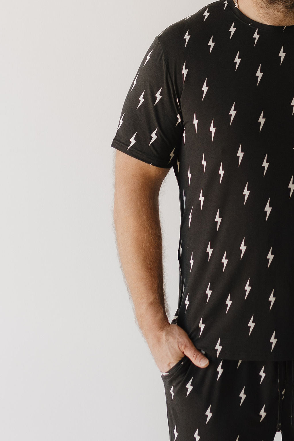A person wearing the Men's Bamboo Short Sleeve Pajamas in Midnight Lightning Bolt by forever french baby stands with one hand in their pocket against a plain, light-colored background, showing only the torso and one arm.