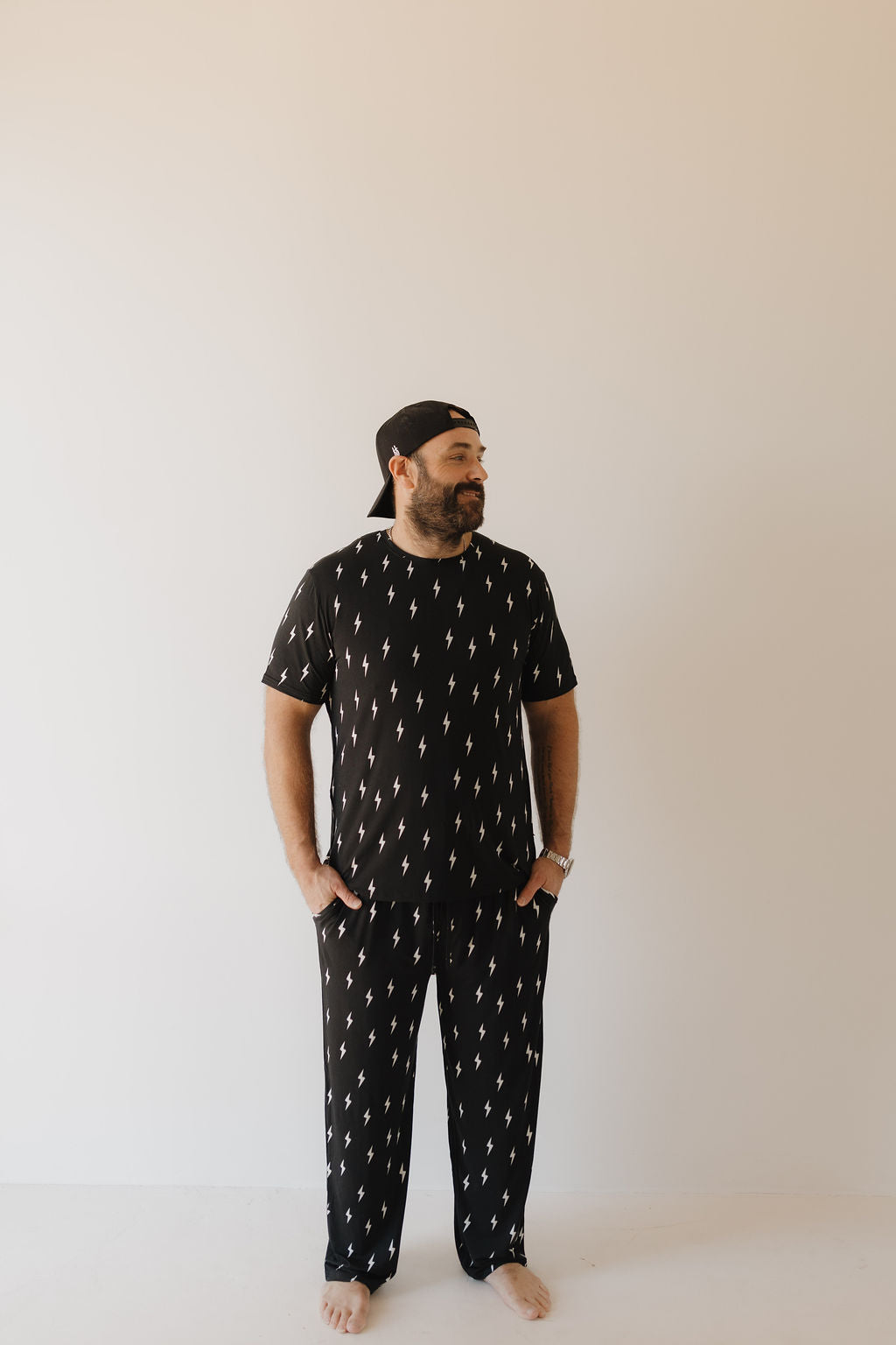 A bearded man stands barefoot against a plain background, wearing Men's Bamboo Short Sleeve Pajamas | Midnight Lightning Bolt by forever french baby. Crafted from breathable fabric, the stylish black ensemble with lightning bolt patterns complements his laid-back look as he wears a backwards cap.
