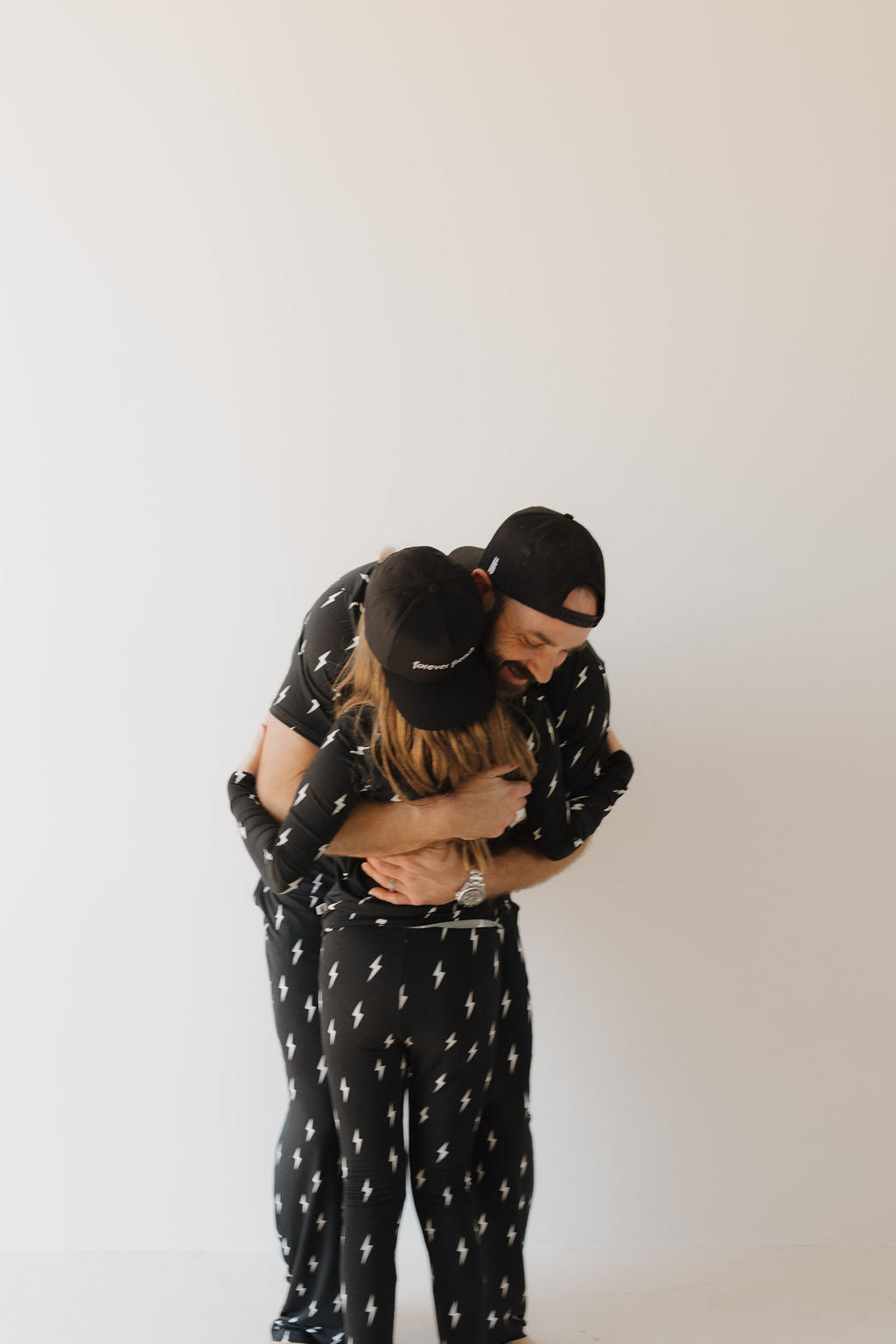 Two people in matching Bamboo Two Piece Pajamas | Midnight Lightning Bolt from forever french baby embrace, their breathable outfit exuding effortless style and comfort. Donning black caps against a plain white wall, they are the epitome of chic relaxation.