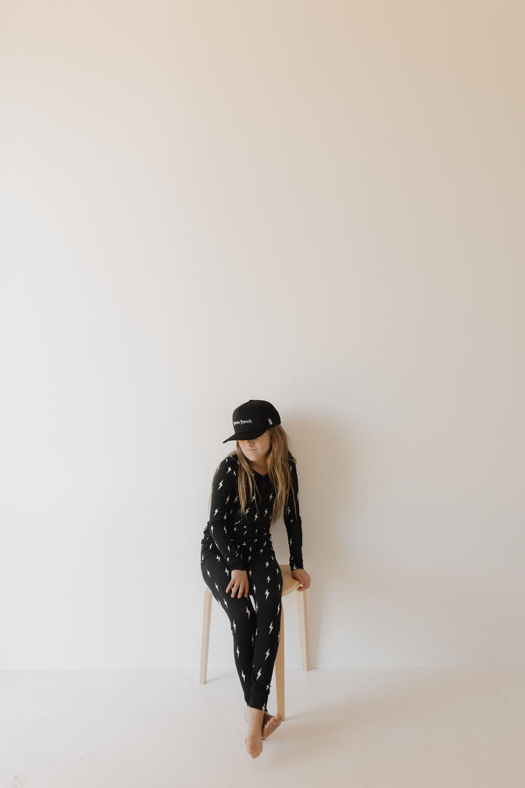 A person sits on a stool against a plain wall, wearing Bamboo Two Piece Pajamas in Midnight Lightning Bolt by forever french baby. Their long hair and relaxed posture complement the minimalistic, bright setting that enhances the tranquil vibe of their sleepwear-inspired outfit.