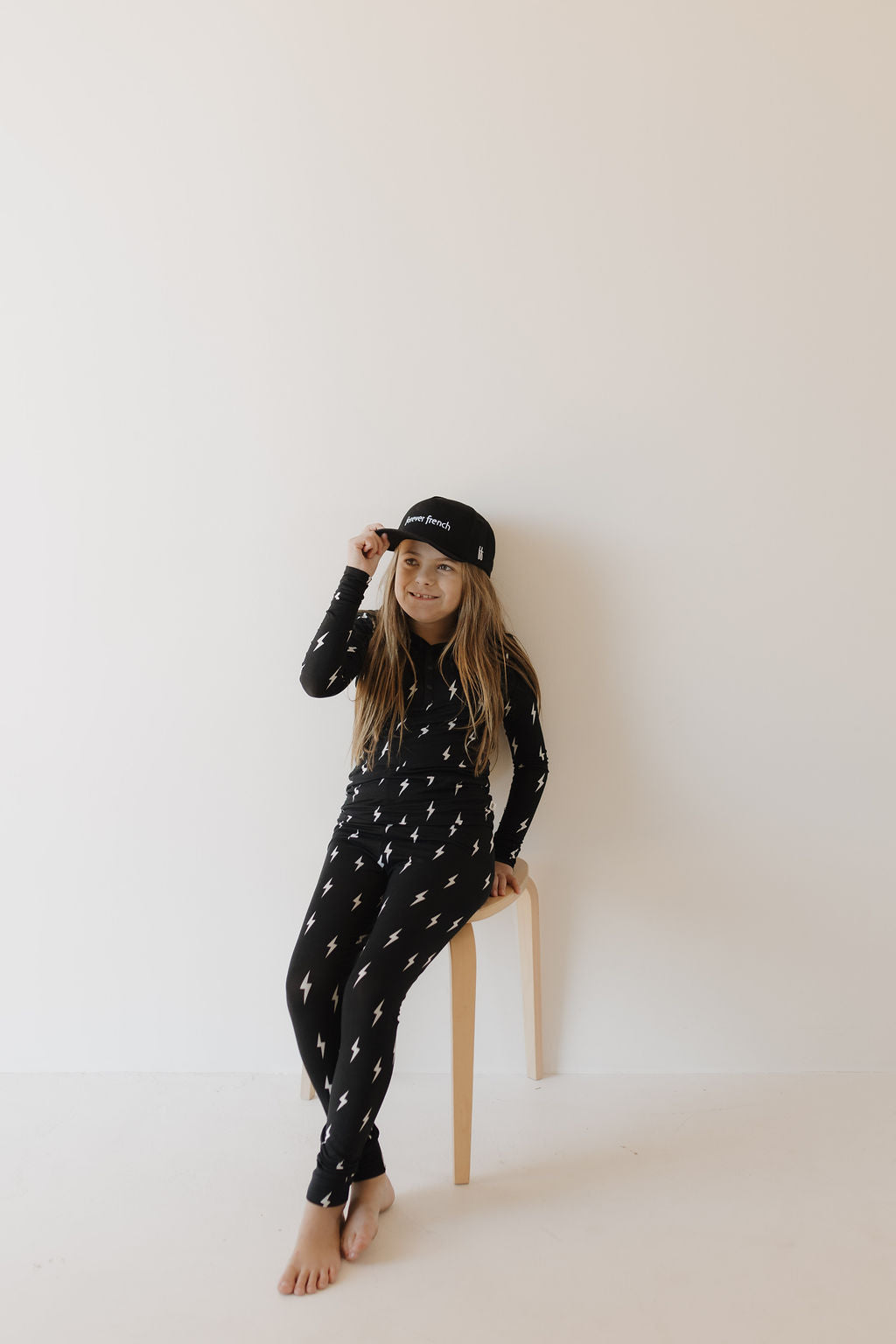 A young girl with long hair sits on a simple wooden stool against a plain background, wearing the Bamboo Two Piece Pajamas in Midnight Lightning Bolt by forever french baby. She is barefoot with a black cap and smiles slightly.