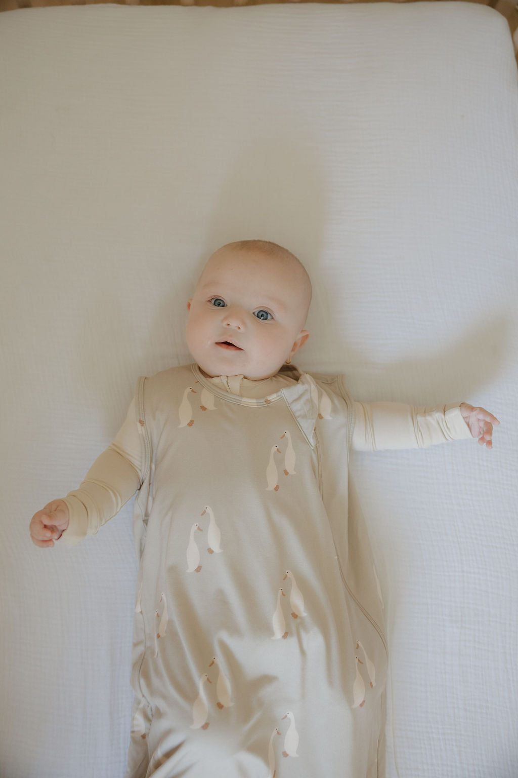 A baby lies on a white sheet, wearing the forever french baby Bamboo Sleep Sack | Silly Goose—a beige bamboo sleep sack with penguin print—looking up adorably with arms outstretched.