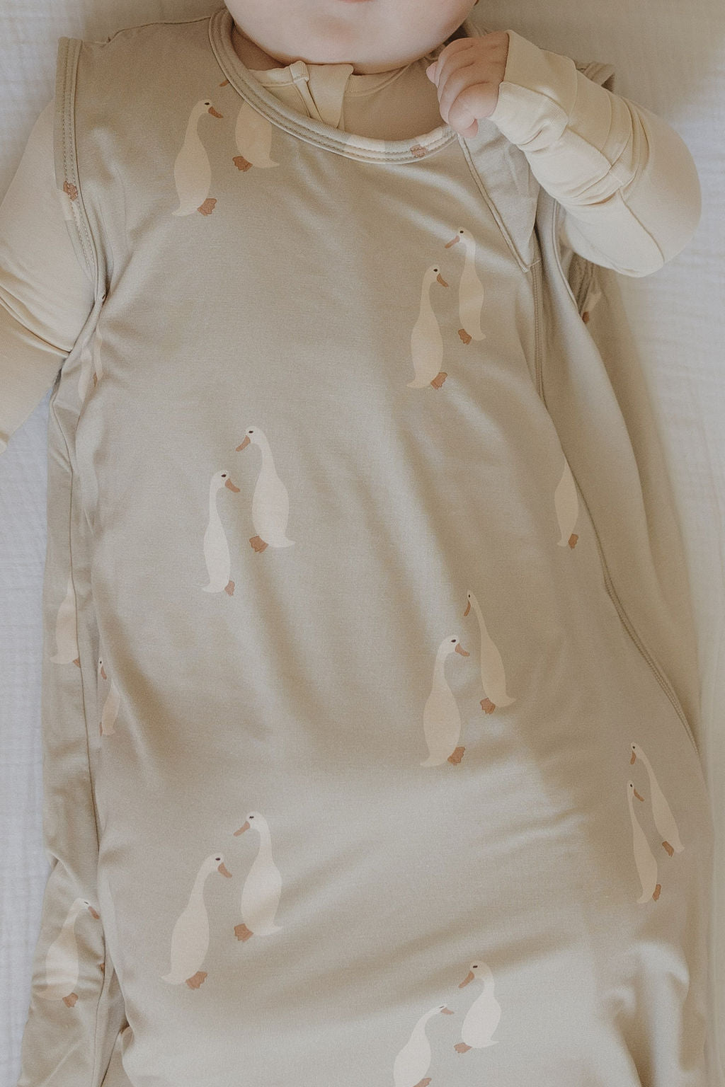A baby wearing the forever french baby Bamboo Sleep Sack | Silly Goose, featuring white duck prints on a cream background, lies on a white surface with only the lower half of their face and one hand visible.