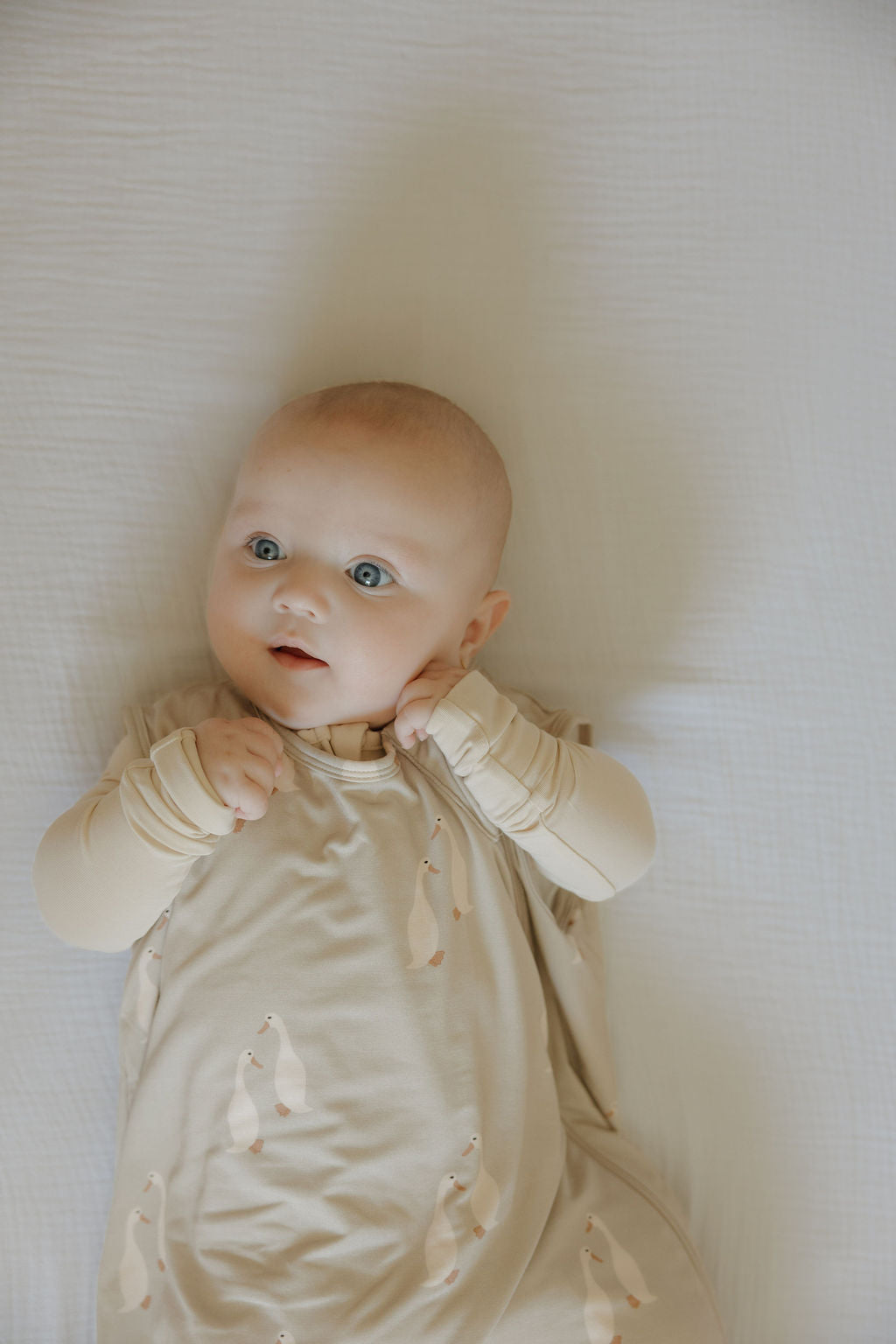 A baby with wide blue eyes lies on a white textured blanket, wearing the forever french baby Bamboo Sleep Sack | Silly Goose and matching long-sleeve mittens decorated with penguin illustrations.
