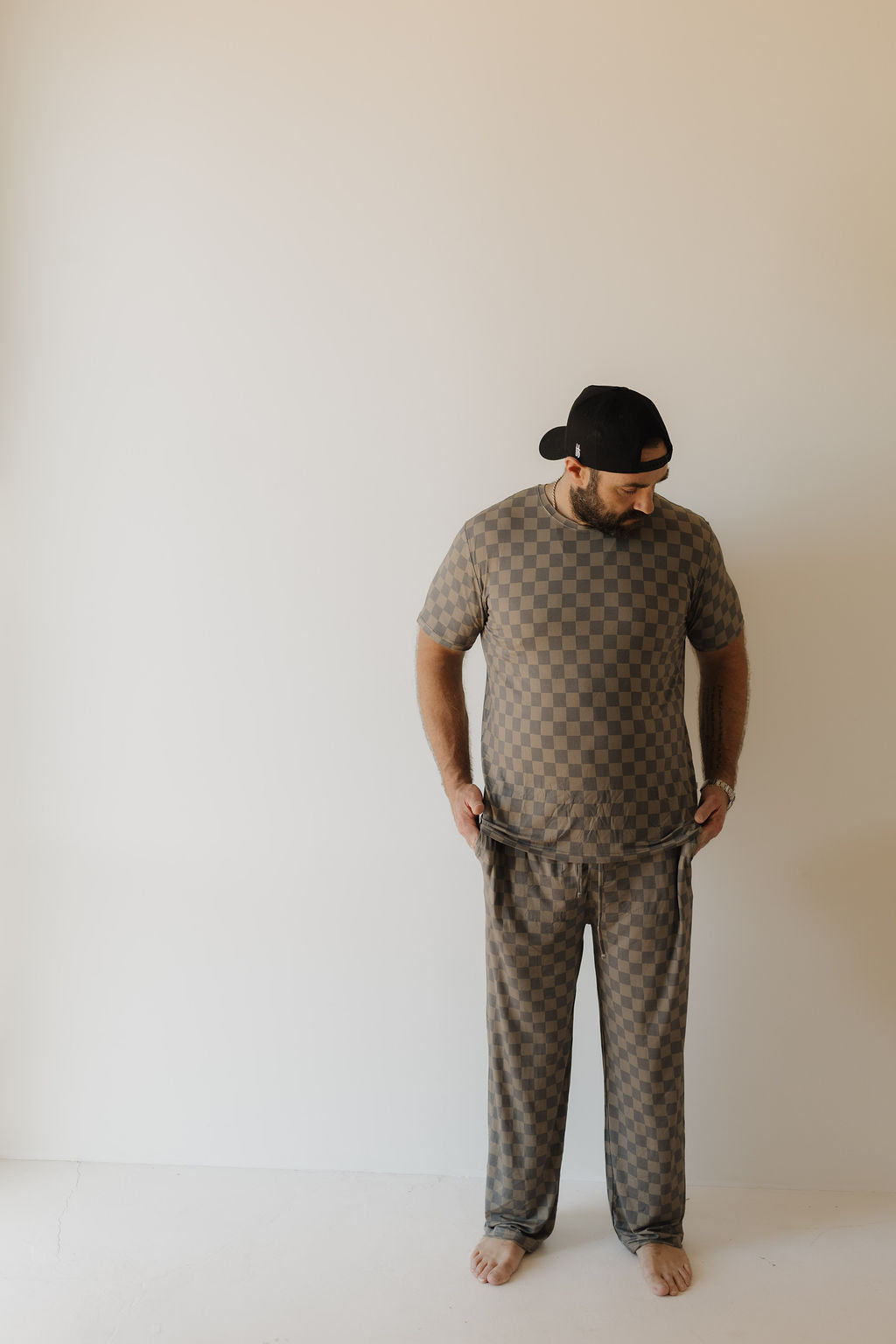 A man stands barefoot against a white wall, wearing forever french baby's Men's Bamboo Short Sleeve Pajama in Faded Checkerboard and a black cap. He looks down with his hands in his pockets.