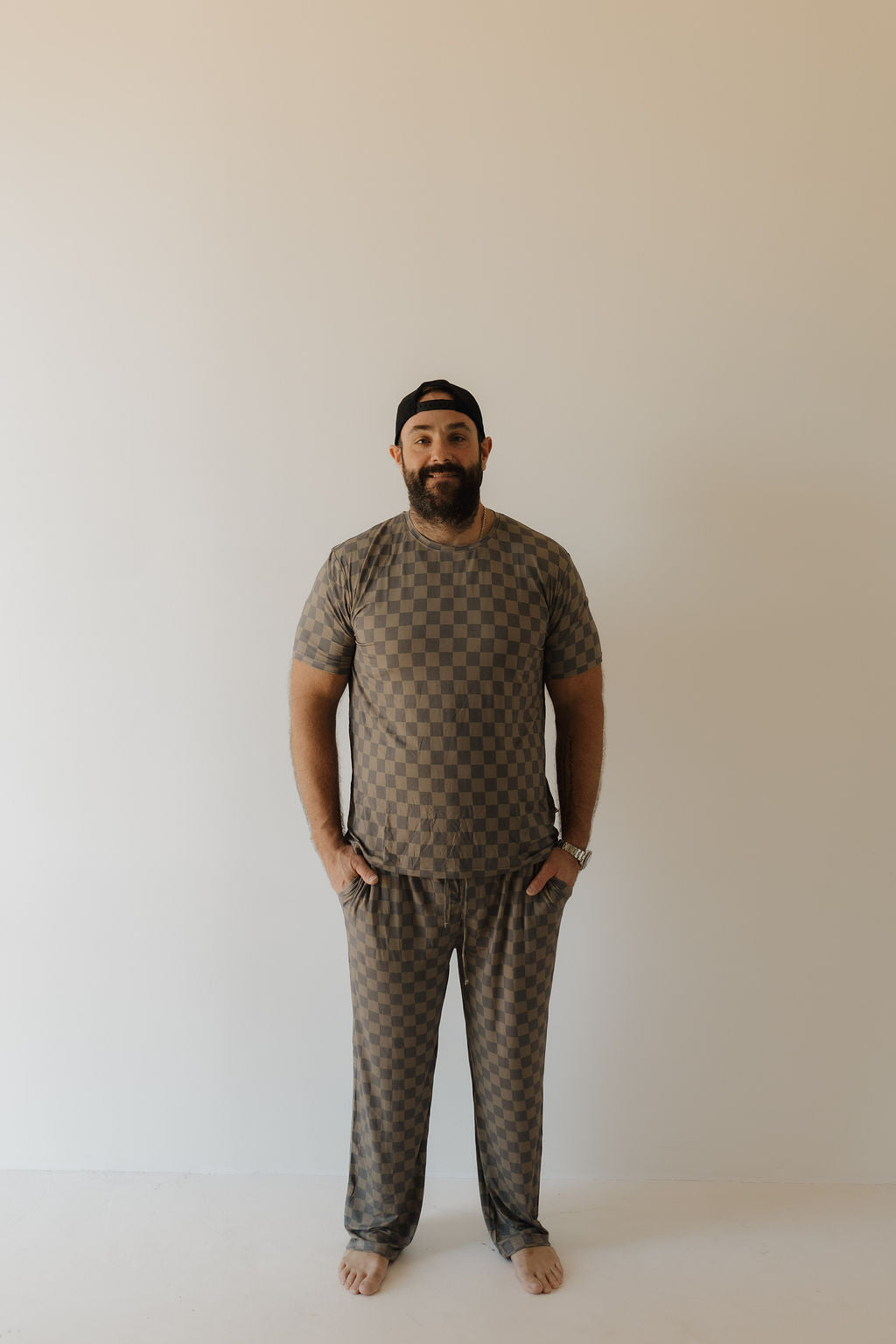 A bearded man stands barefoot on a plain background in forever french baby's Men's Bamboo Short Sleeve Pajama in Faded Checkerboard. With hands in pockets and a black cap, he exudes casual elegance and style, wearing hypoallergenic sleepwear that balances comfort with flair.