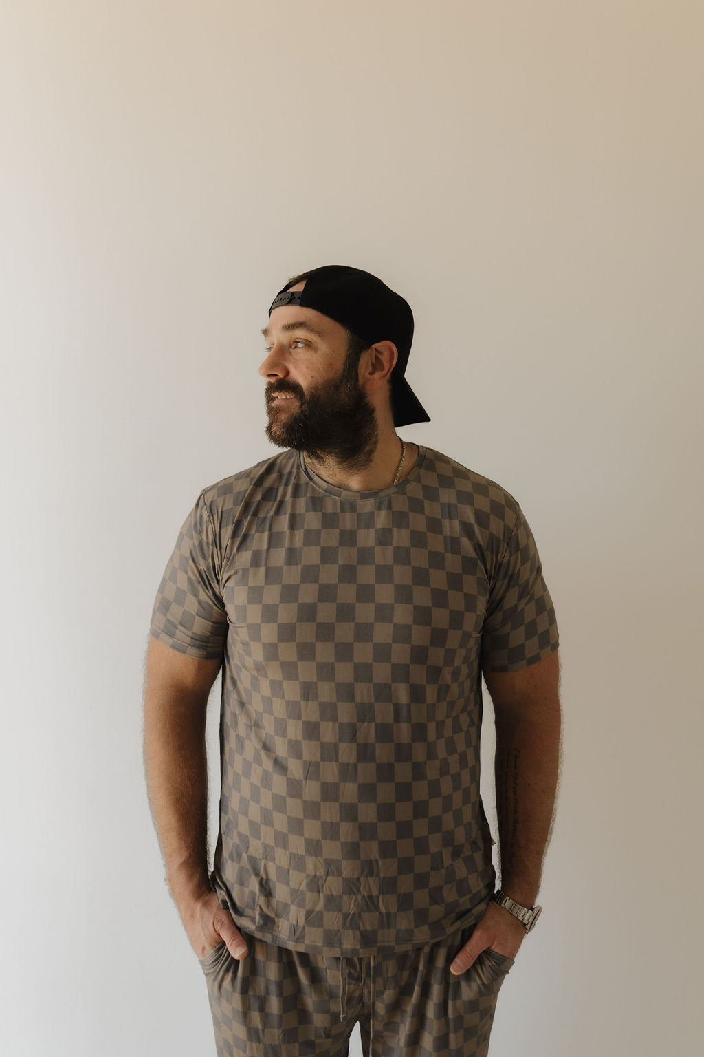 A man in Forever French Baby's Men's Bamboo Short Sleeve Pajama, featuring a Faded Checkerboard design, stands on a plain backdrop. With hands in pockets and wearing a backwards baseball cap, he gazes sideways.