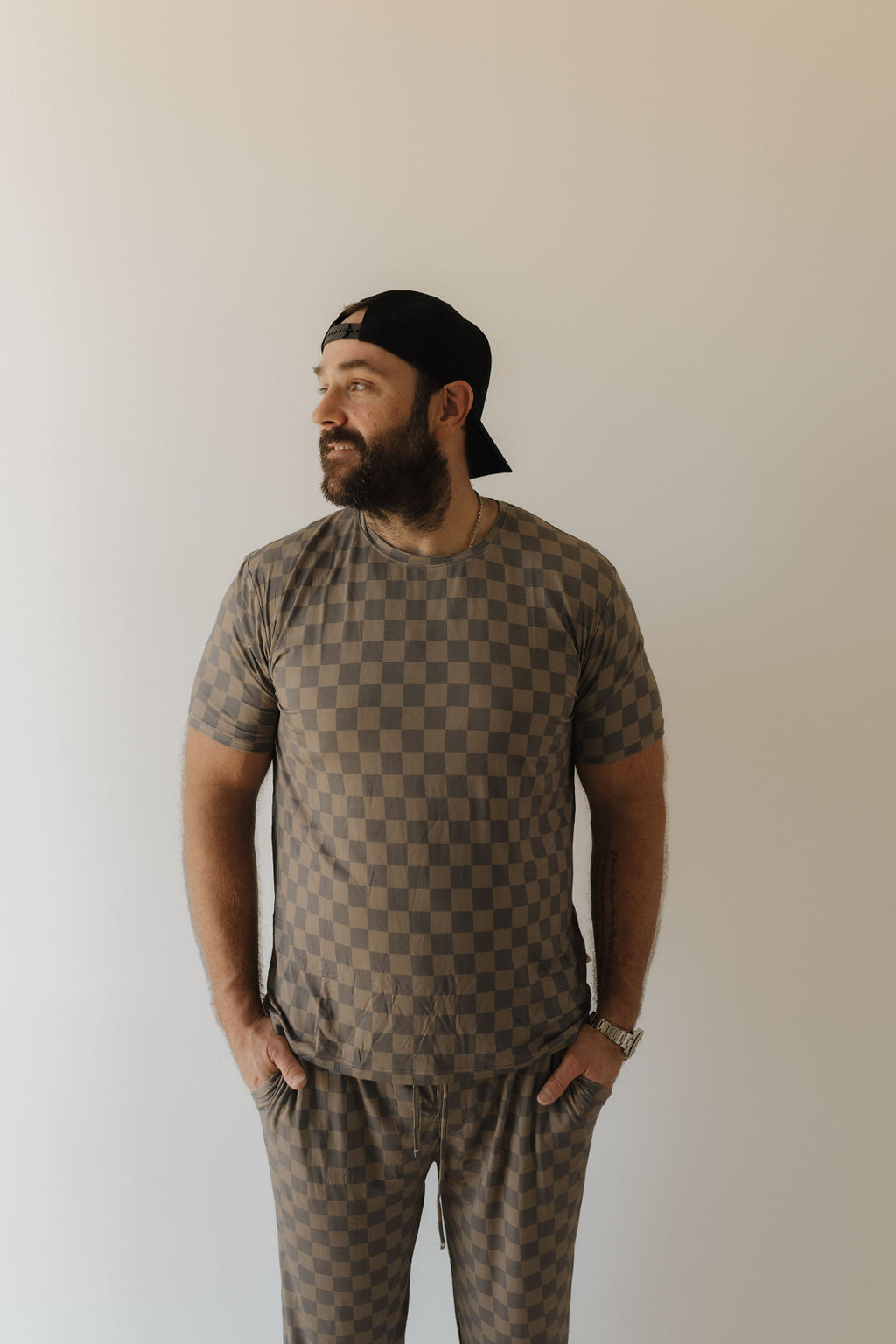 A bearded man in a black cap, facing left, wears a brown checkered Men's Bamboo Short Sleeve Pajama by forever french baby. Standing against a plain light background, he has his hands in his pockets.