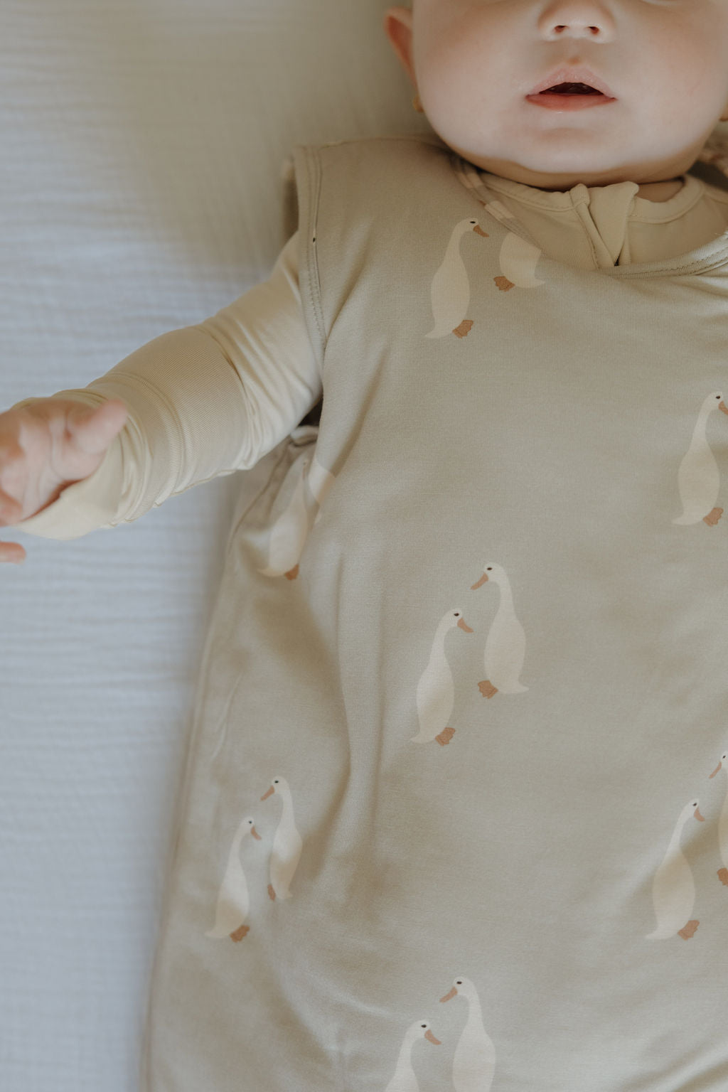 A baby wearing the forever french baby Bamboo Sleep Sack | Silly Goose, featuring a duck pattern, lies on a light surface. Only the lower half of the baby's face and one arm are visible, emphasizing the cozy wearable blanket.