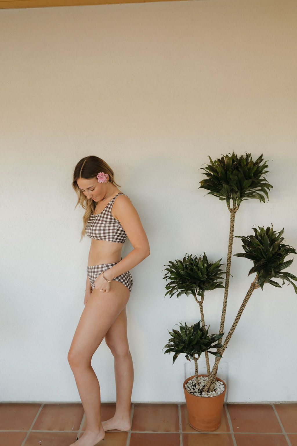A model poses in profile wearing the Women's Two Piece Swimsuit in Chocolate Gingham by forever french baby, exuding charm with a flower in their hair and barefoot on tiled flooring. A potted plant enhances the Forever French Baby vibe beside them.