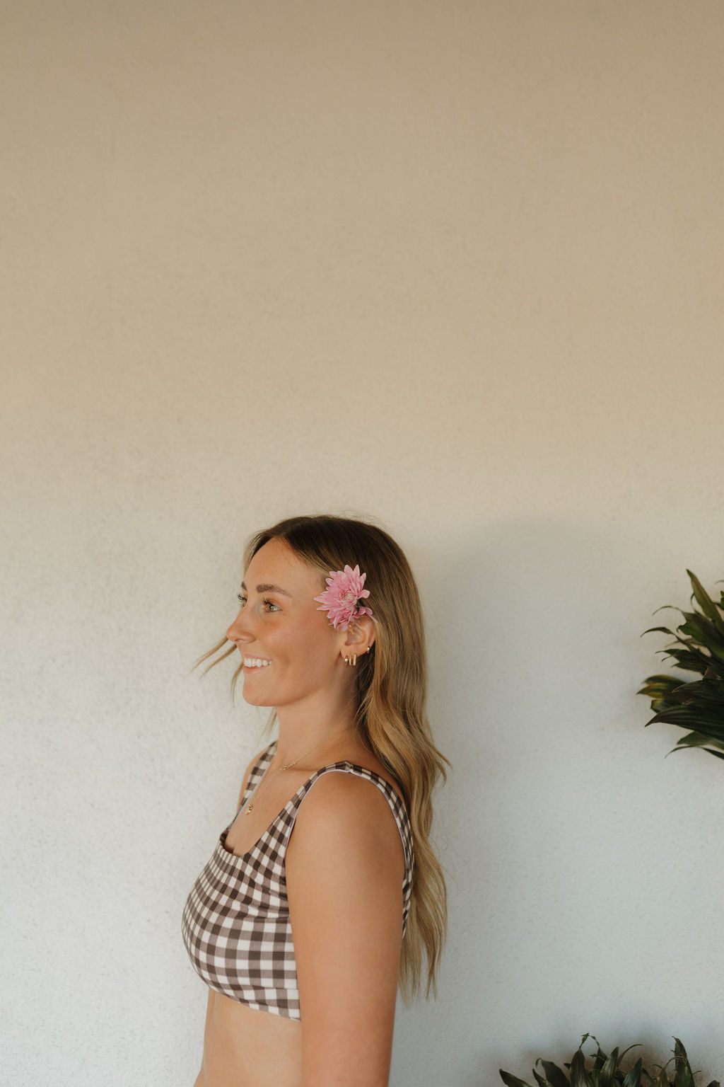 A person with long hair stands in profile, wearing a Women's Two Piece Swimsuit from the Chocolate Gingham collection by forever french baby, a pink flower behind their ear. They're smiling against a neutral wall with a green plant visible on the side.