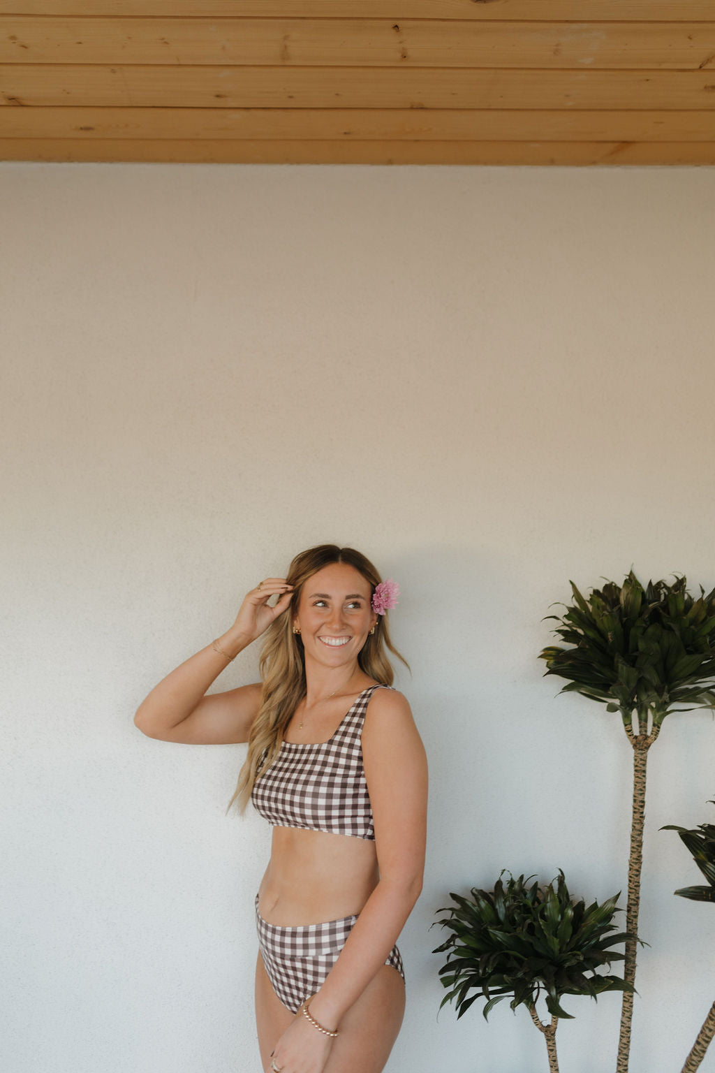 A person with long hair wears a Women's Two Piece Swimsuit in Chocolate Gingham from forever french baby, smiling with a pink flower near their ear. They pose by a plant against a white wall and wooden ceiling, showcasing the brand's essence.