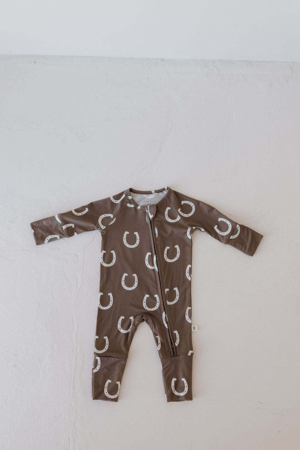 The Bamboo Zip Pajamas | Giddy Up by forever french baby is a brown jumpsuit with white horseshoes, resting flat on a light gray surface. Made from breathable fabric, it has a front zipper and long sleeves for comfort.