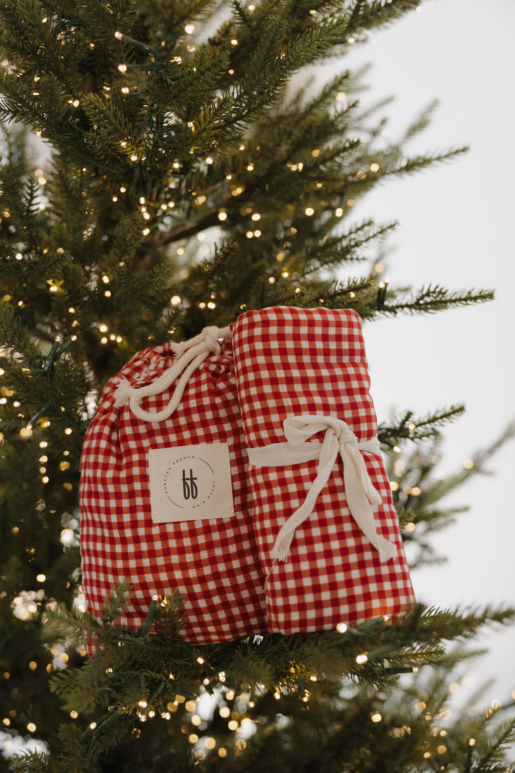 Two Muslin Swaddle | Red Gingham by forever french baby hang on a Christmas tree with warm white lights, adding festive charm.