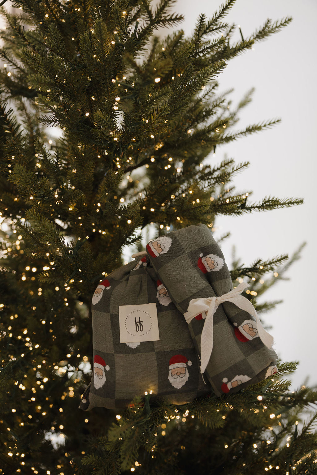 Two Muslin Swaddle | Santa Checkerboard wraps by forever french baby add a cozy, festive glow as they hang like gift bags on a Christmas tree decorated with warm white string lights and lush, green branches.