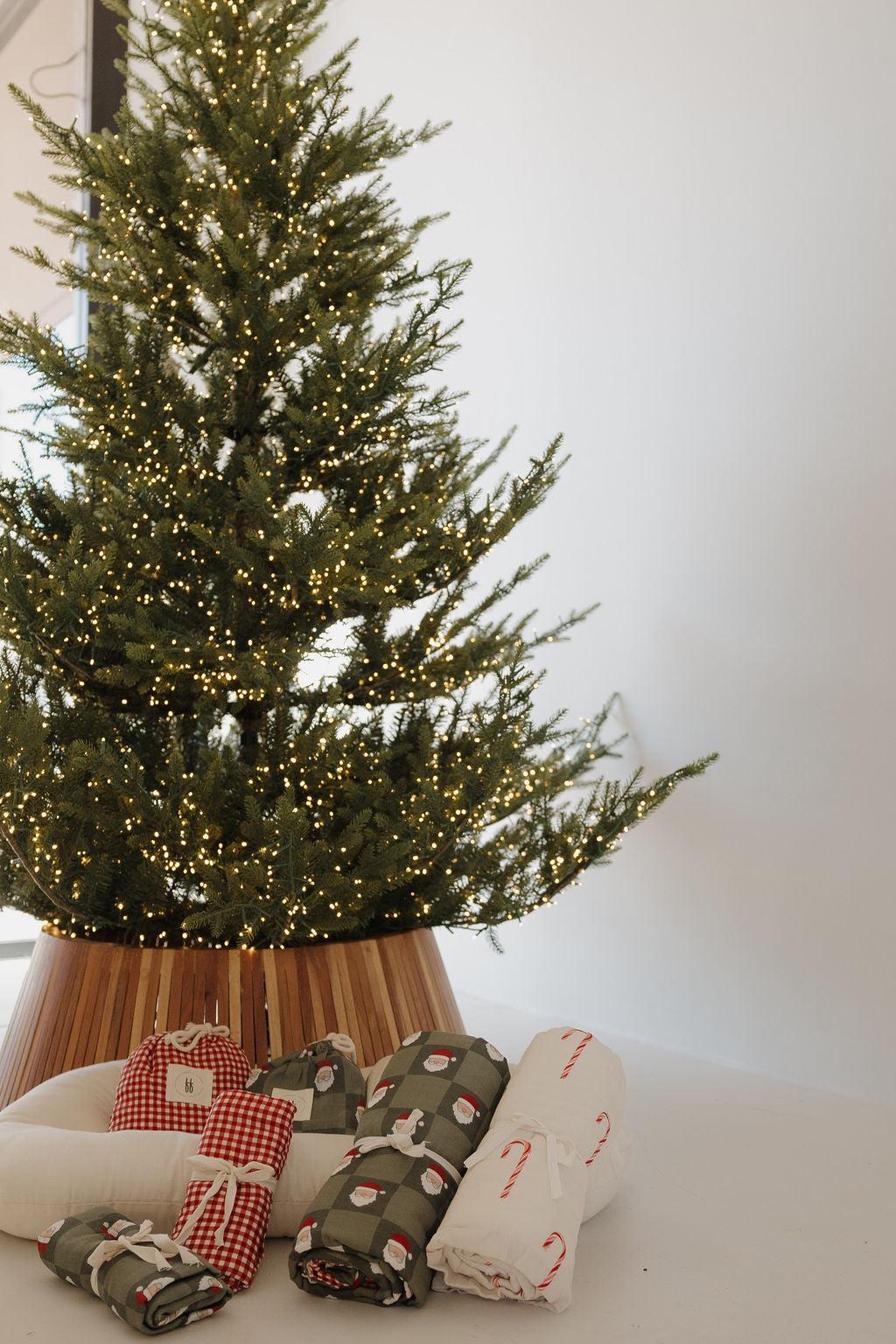 A decorated Christmas tree with white lights stands in a wooden collar. In front are folded blankets, festive pillows, and the Muslin Crib Sheet | Candy Cane (Preview) by forever french baby—perfect for adding holiday cheer to your nursery.