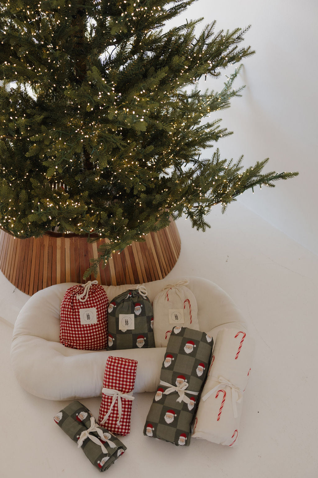 A Christmas tree with white lights stands above gifts, including the forever french baby Muslin Swaddle | Santa Checkerboard, wrapped in festive paper with Santa faces, candy canes, and red gingham checks.