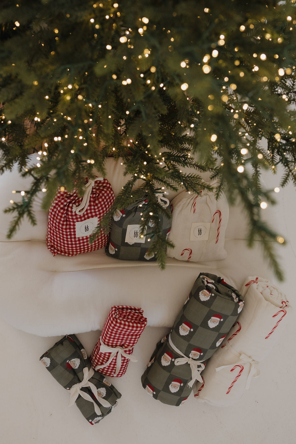 An overhead view of wrapped gifts under a festive tree, including the forever french baby Muslin Quilt | Candy Cane (Preview) in red, green, and cream with Santa faces and candy canes. Warm fairy lights twinkle above.