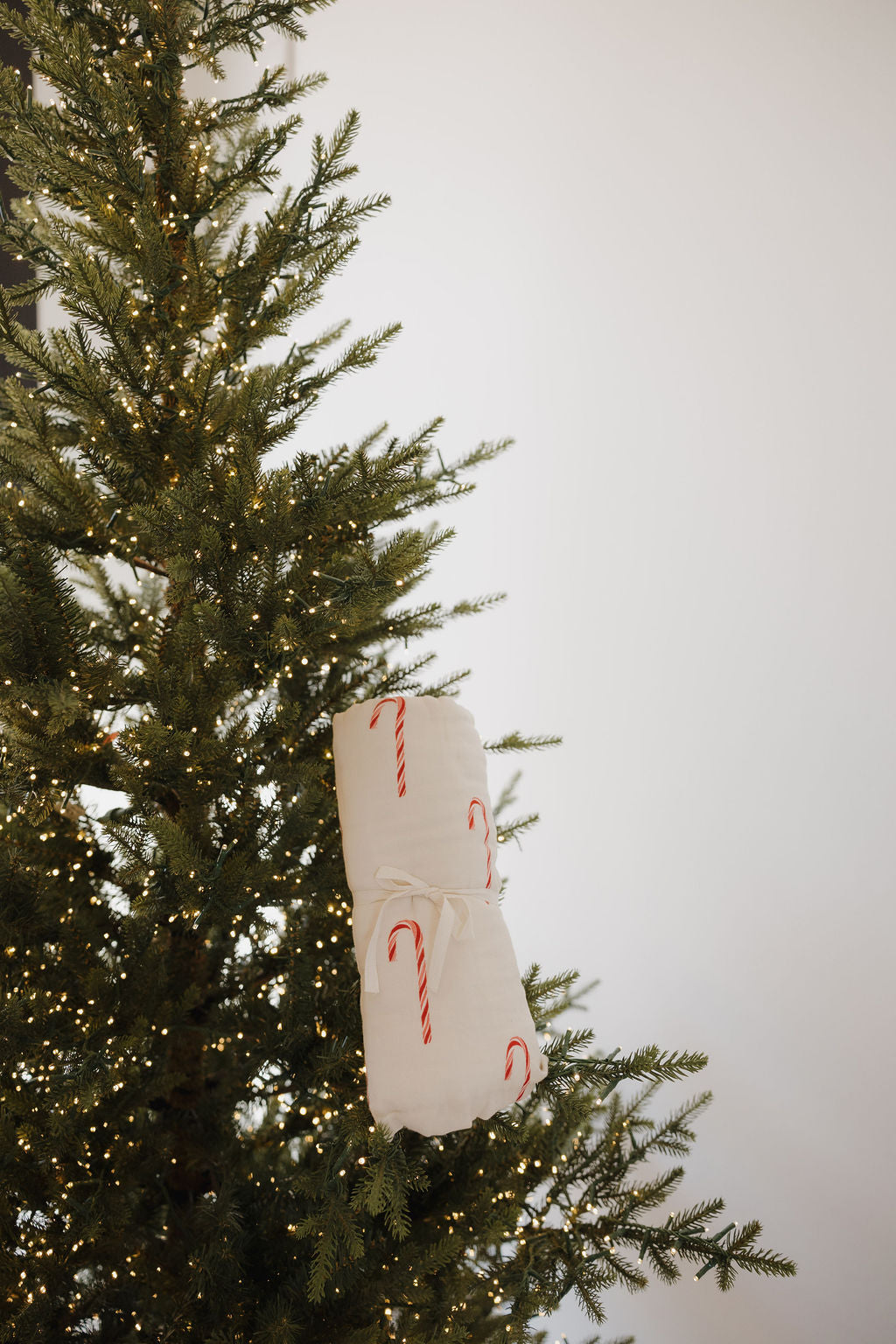 A decorated Christmas tree with warm white lights holds the forever french baby Muslin Quilt | Candy Cane (Preview), rolled up and tied with string, adorned with three candy canes, against a plain white background.