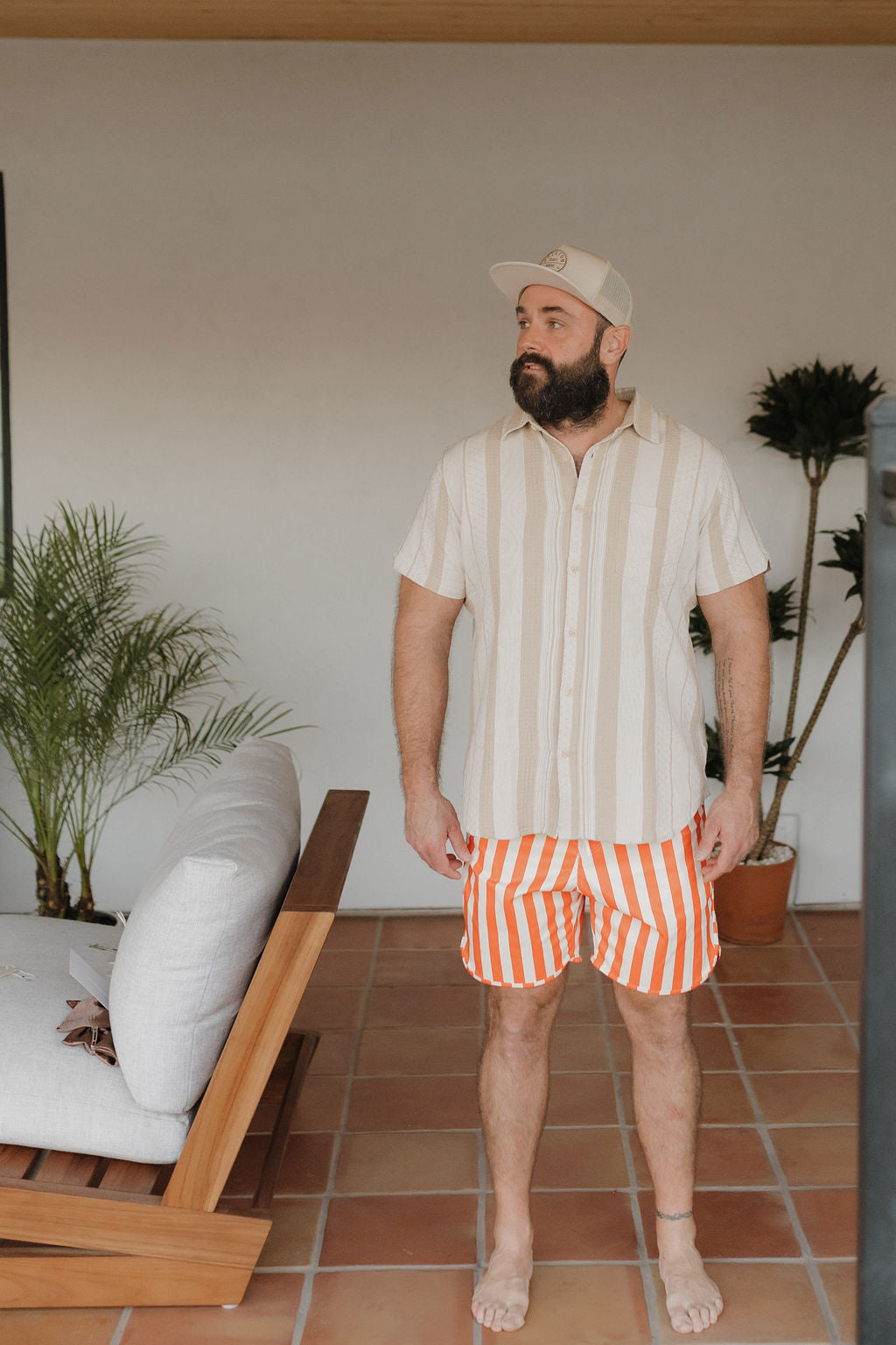 A bearded man stands on a tiled floor, wearing an off-white cap and striped short-sleeve shirt, stylishly paired with orange and white Men's Boardshort from forever french baby's Poolside Collection. A wooden sofa with white cushions and lush potted plants are in the background.