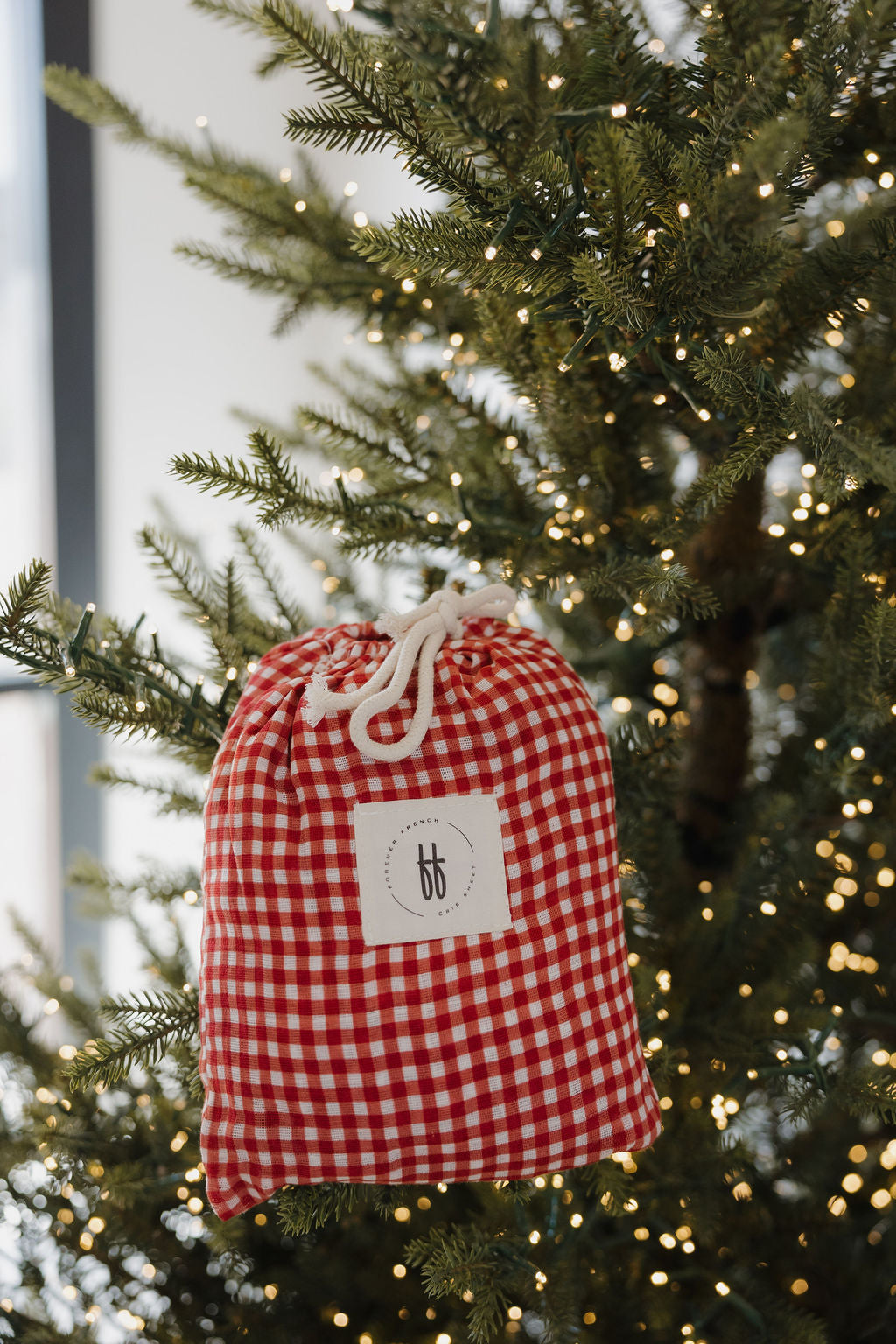 A red and white checkered drawstring bag hangs on a Christmas tree with warm white lights, echoing the cozy look of the Muslin Crib Sheet | Red Gingham by forever french baby.