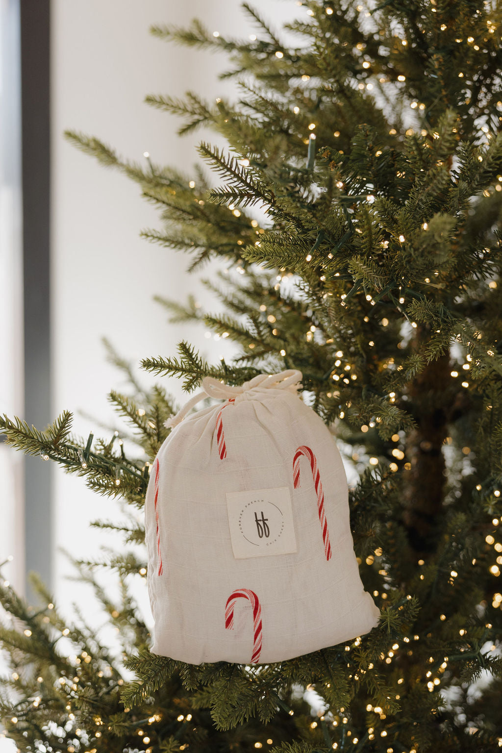 A white cloth gift bag with candy canes hangs on a Christmas tree with warm lights, reflecting the festive style of the forever french baby Muslin Crib Sheet | Candy Cane (Preview)—a charming choice for your nursery bedding.