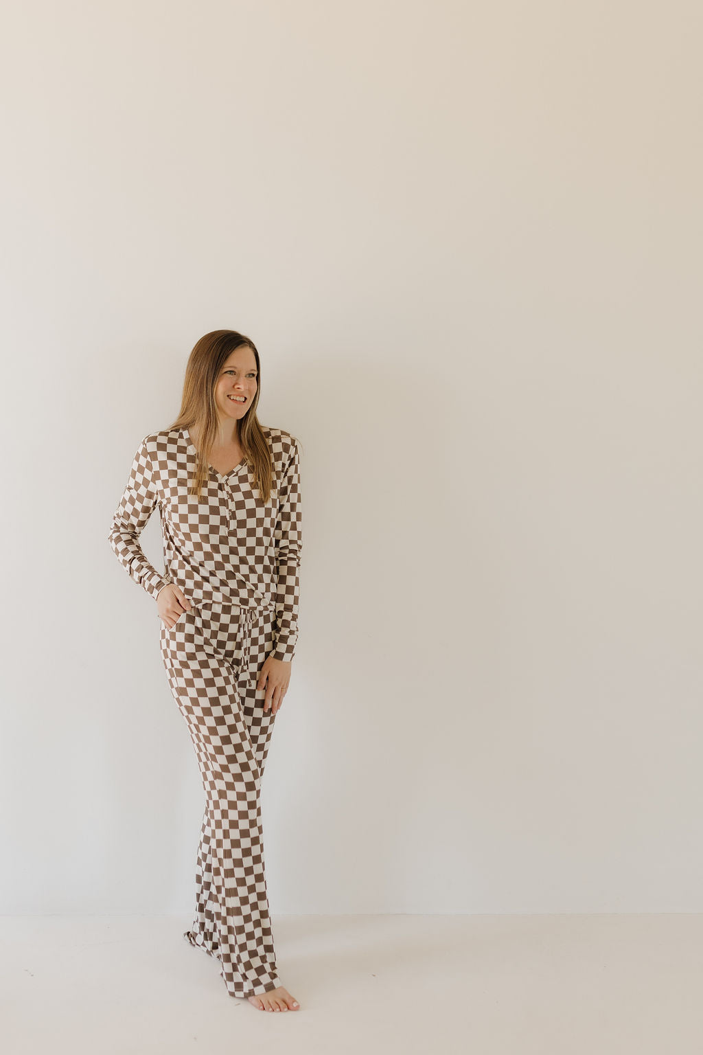 A person stands barefoot against a plain background, wearing forever french baby Women's Bamboo Pajama | Dutton in a brown and white checkerboard pattern. The hypo-allergenic bamboo fabric complements their smile and long hair perfectly.