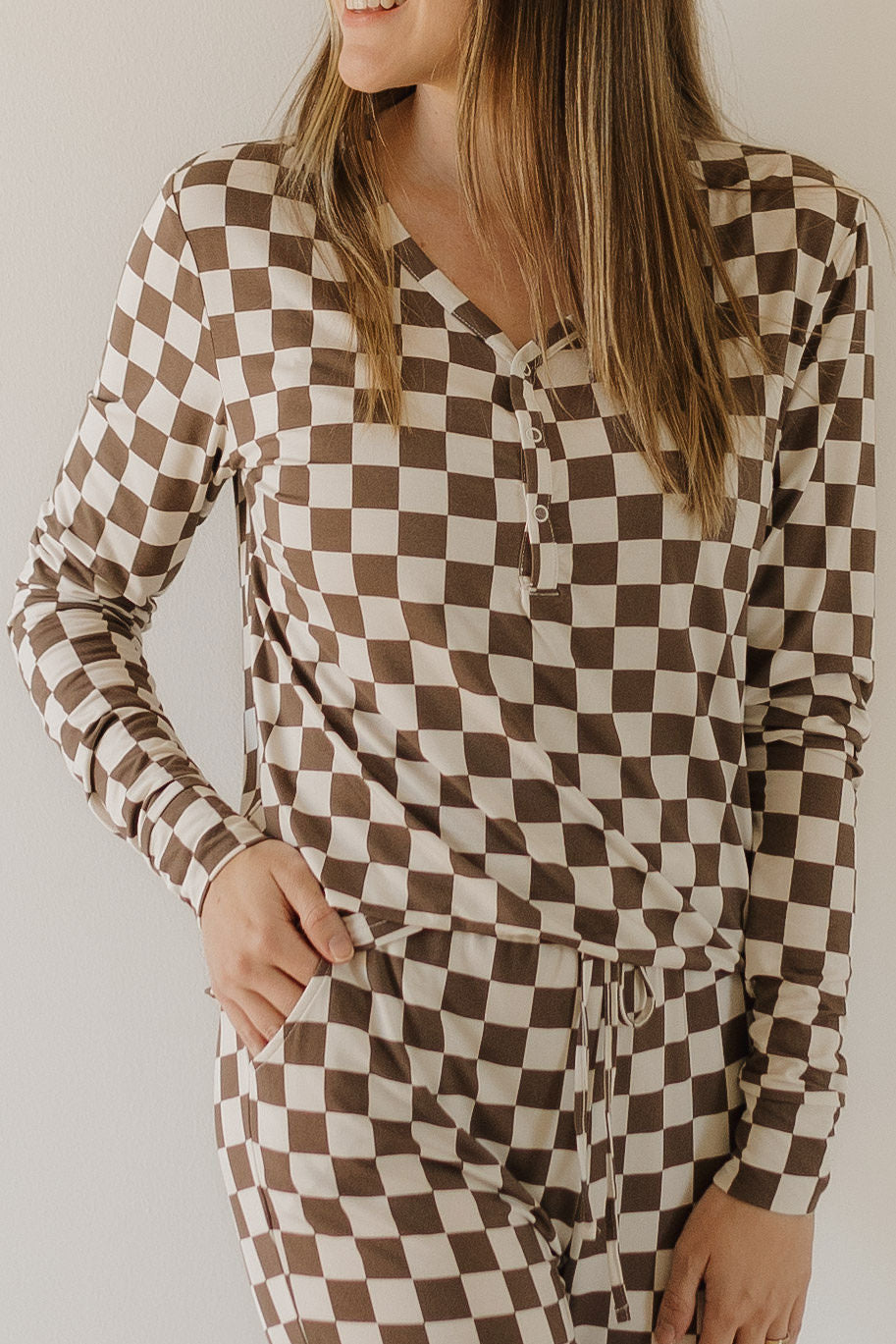 In a white backdrop, a woman wears the Women's Bamboo Pajama by forever french baby. This hypo-allergenic ensemble features a brown and white checkered pattern with buttons at the neckline, ensuring comfort with breathable bamboo fabric as she smiles with one hand on her hip.