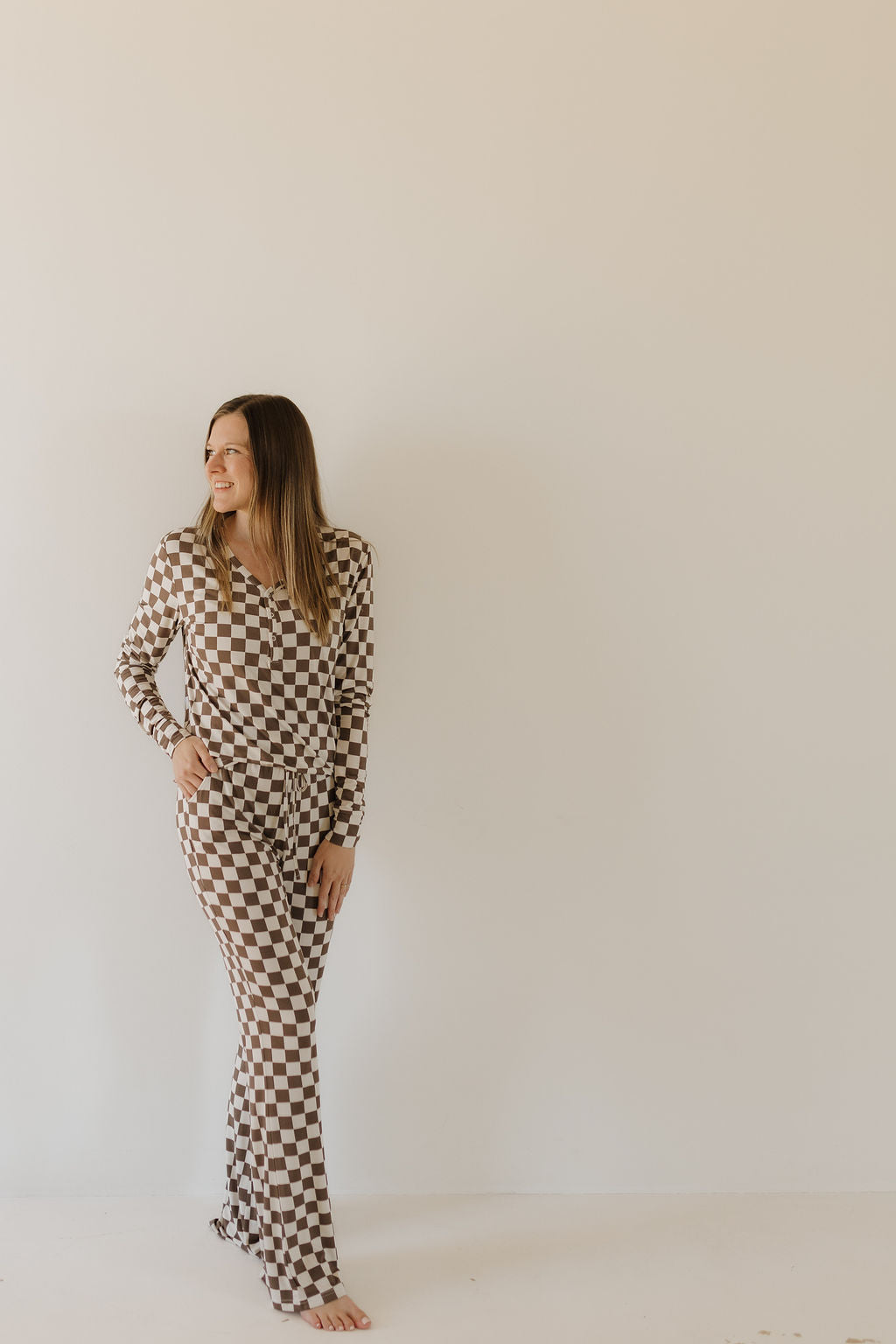 A woman smiles, looking to the side against a plain background. She wears forever french baby's Women's Bamboo Pajama | Dutton, a long-sleeve, brown and white checkered jumpsuit crafted from breathable bamboo fabric, appearing relaxed with one hand in her pocket.