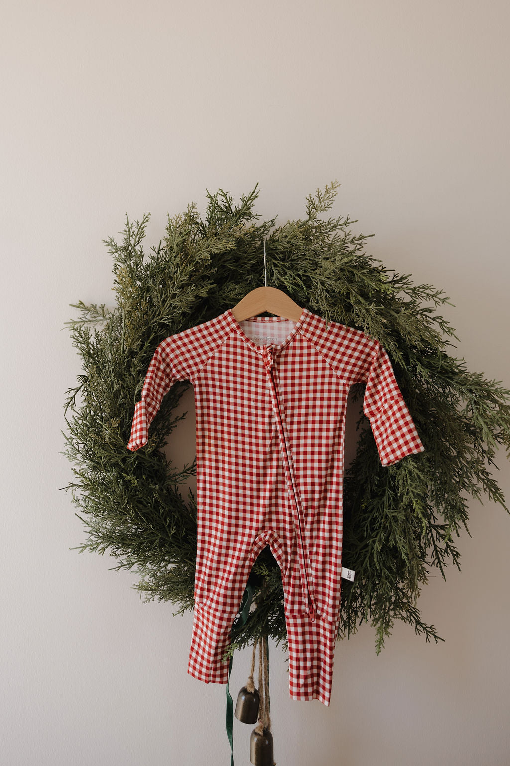 The Bamboo Zip Pajamas in Red Gingham by forever french baby hang on a wooden hanger, displayed before a green holiday wreath against a light-colored wall.
