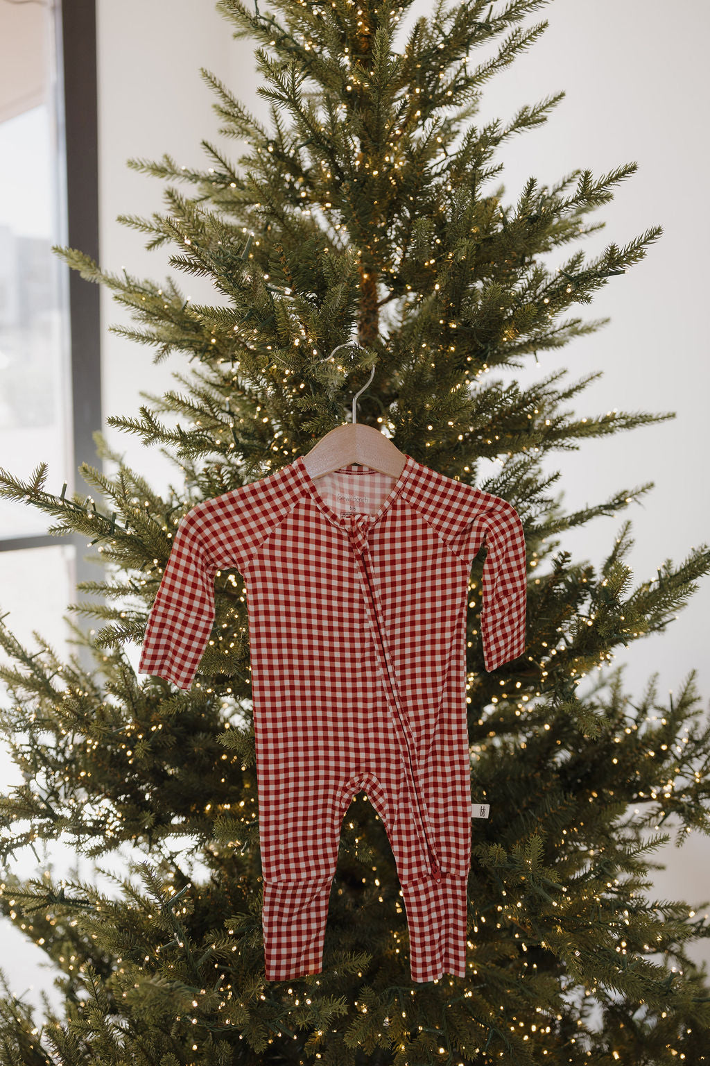 A forever french baby Bamboo Zip Pajamas in red gingham hangs on a wooden hanger in front of a Christmas tree adorned with twinkling white lights.