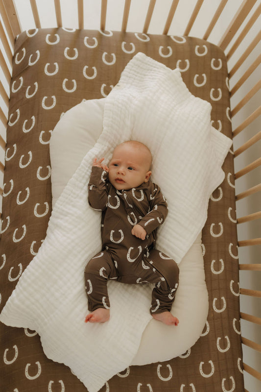 A baby lies in a crib on a brown sheet with white horseshoe patterns, wearing Bamboo Zip Pajamas | Giddy Up by forever french baby. The child rests on a white pillow and blanket, while the wooden slat crib ensures comfort and style with its breathable fabric ensemble.