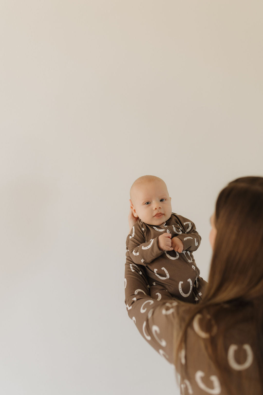 A person holds a baby in forever french baby's Bamboo Zip Pajamas, Giddy Up design, featuring a white horseshoe pattern on brown. The individual, viewed from behind, is also dressed in matching breathable fabric attire against a plain light-colored wall.