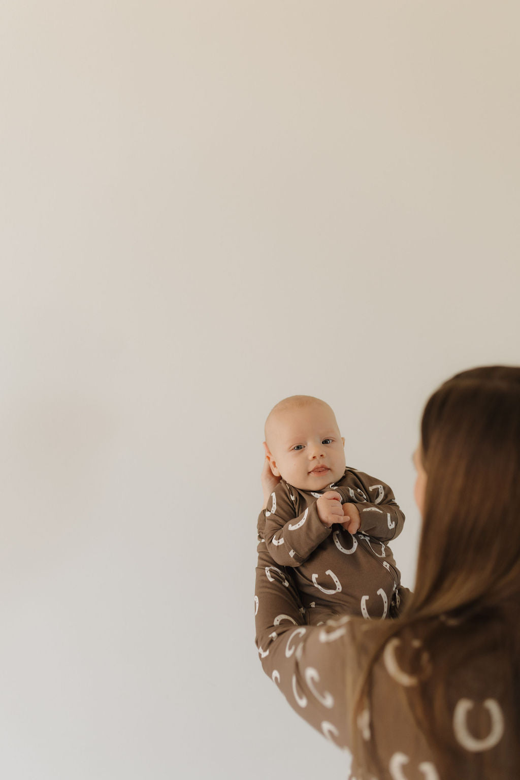 A person with long brown hair holds a baby in Bamboo Zip Pajamas | Giddy Up by forever french baby. The baby, dressed in breathable fabric with white horseshoe patterns, looks directly at the camera against a plain, light-colored wall.