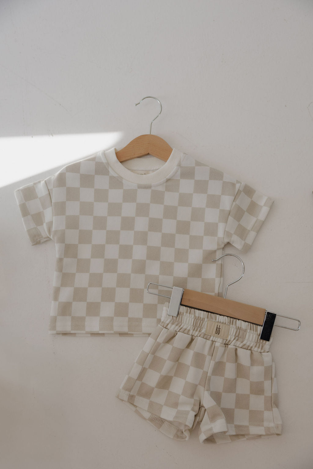 The French Terry Short Set | Checkerboard by forever french baby, featuring a beige and white checkered short sleeve top and matching shorts, hangs on wooden hangers against a light-colored wall.