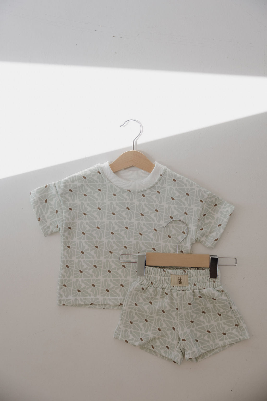 The French Terry Short Set | Coast by forever french baby, featuring a pale green geometric pattern with brown floral accents and subtle FF logo details, hangs on a wooden hanger against a white wall.