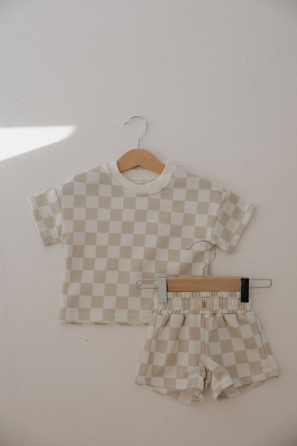 The forever french baby French Terry Short Set | Checkerboard features a beige and white checkered short sleeve top and matching shorts for children, both displayed on wooden hangers against a light-colored wall.