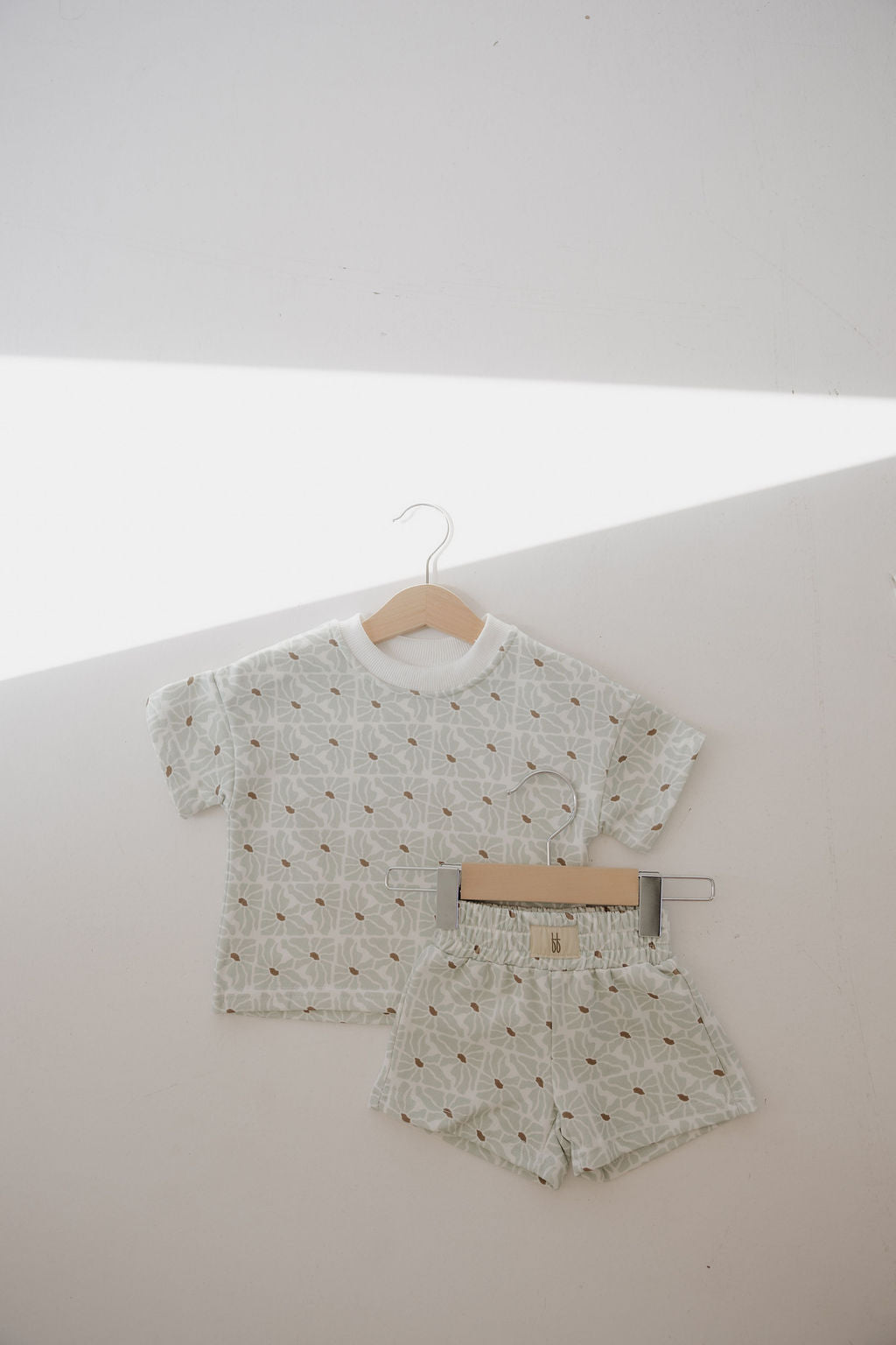 The forever french baby French Terry Short Set | Coast in light green, featuring a subtle geometric pattern, hangs on a wooden hanger with the matching shorts clipped below, bright sunlight casting diagonal shadows across the scene.