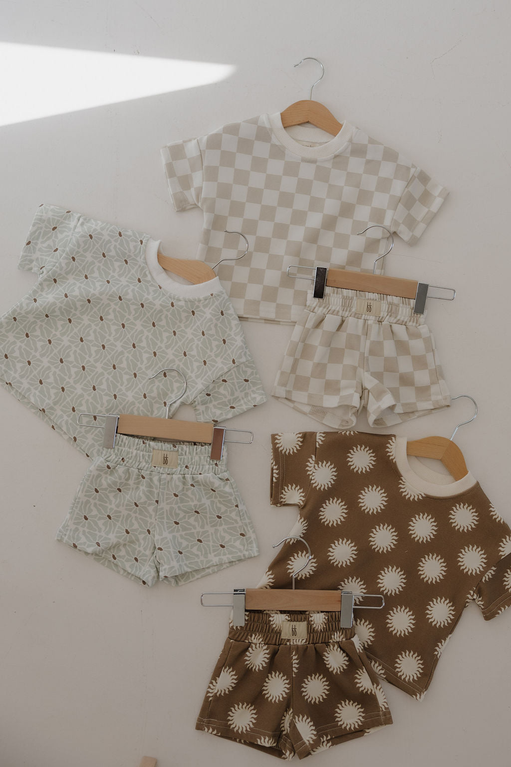 Three French Terry Short Sets by forever french baby hang on display. Each Coast set features kids’ matching shorts and soft short sleeve tops in beige checkerboard, pale green geometric, and brown with white sunburst patterns.