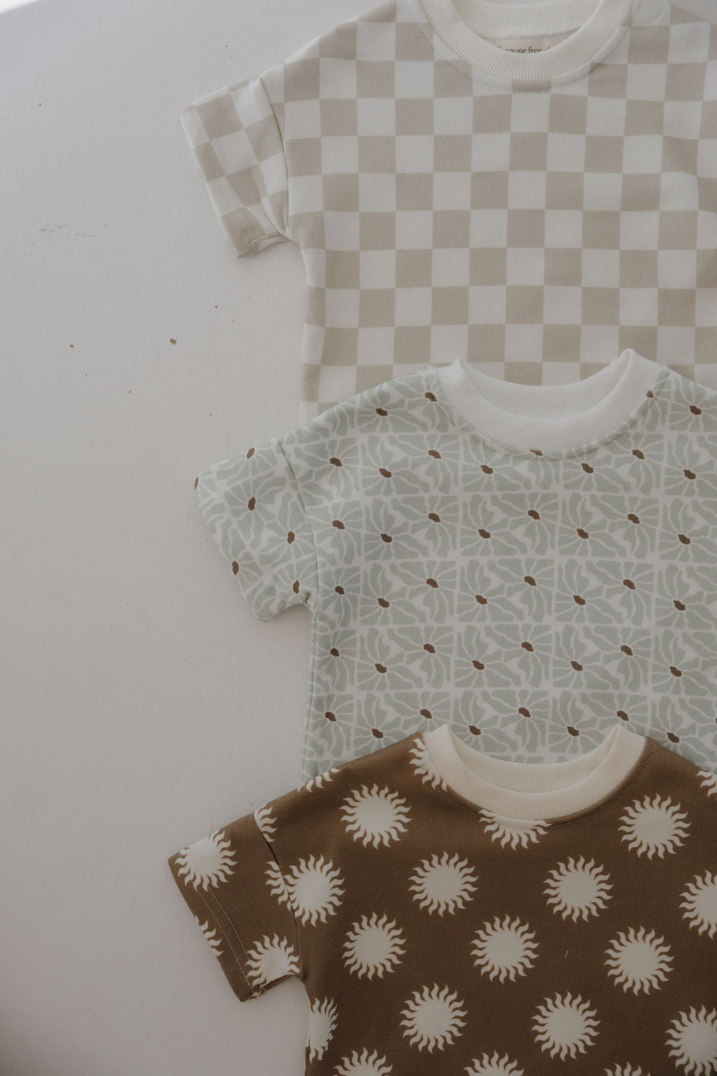 Three short-sleeve baby shirts from forever french baby’s French Terry Short Set—Coast: beige/white checkerboard, pale blue geometric, and brown with white sunbursts—are shown laid flat.