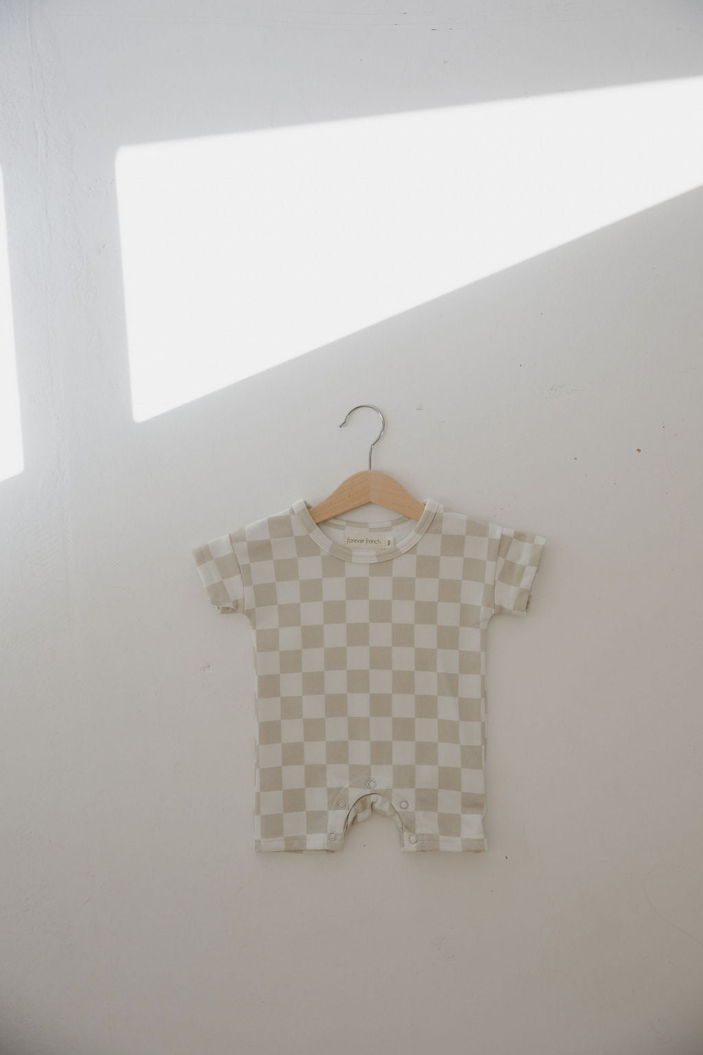 The forever french baby French Terry Romper | Checkerboard, featuring a beige and white check pattern and handy snap closures, hangs on a wooden hanger against a light wall bathed in sunlight—a charming baby essential.