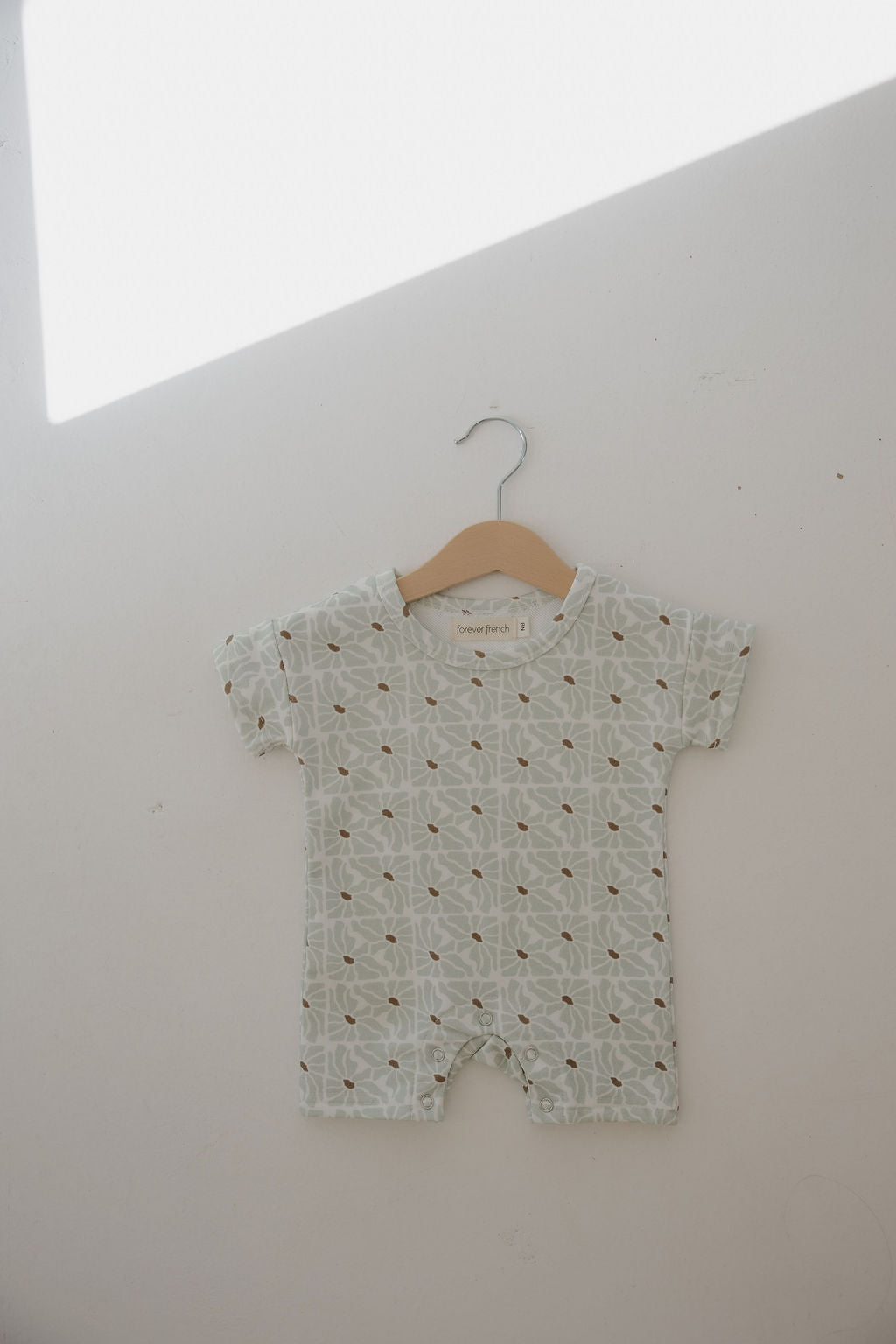 The forever french baby French Terry Romper | Coast, a short-sleeved, light-colored romper with a leafy pattern, hangs on a wooden hanger against a white wall—ideal breathable apparel for newborns on warm days.