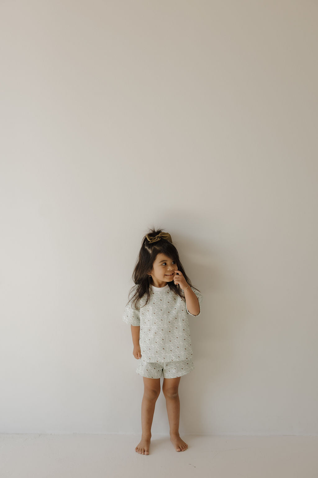 A young girl with long dark hair stands barefoot against a light wall, wearing the forever french baby French Terry Short Set | Coast. She looks to the side and touches her face with one hand.