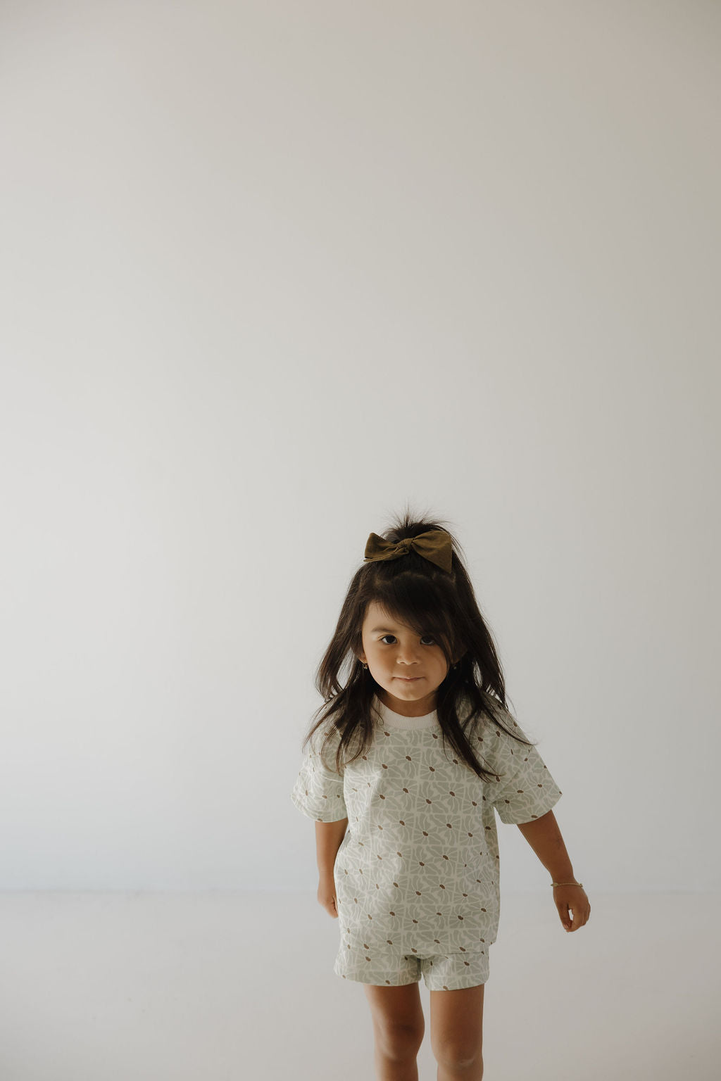 A young girl with long dark hair and a bow stands in a bright, minimal room, wearing the forever french baby French Terry Short Set | Coast.
